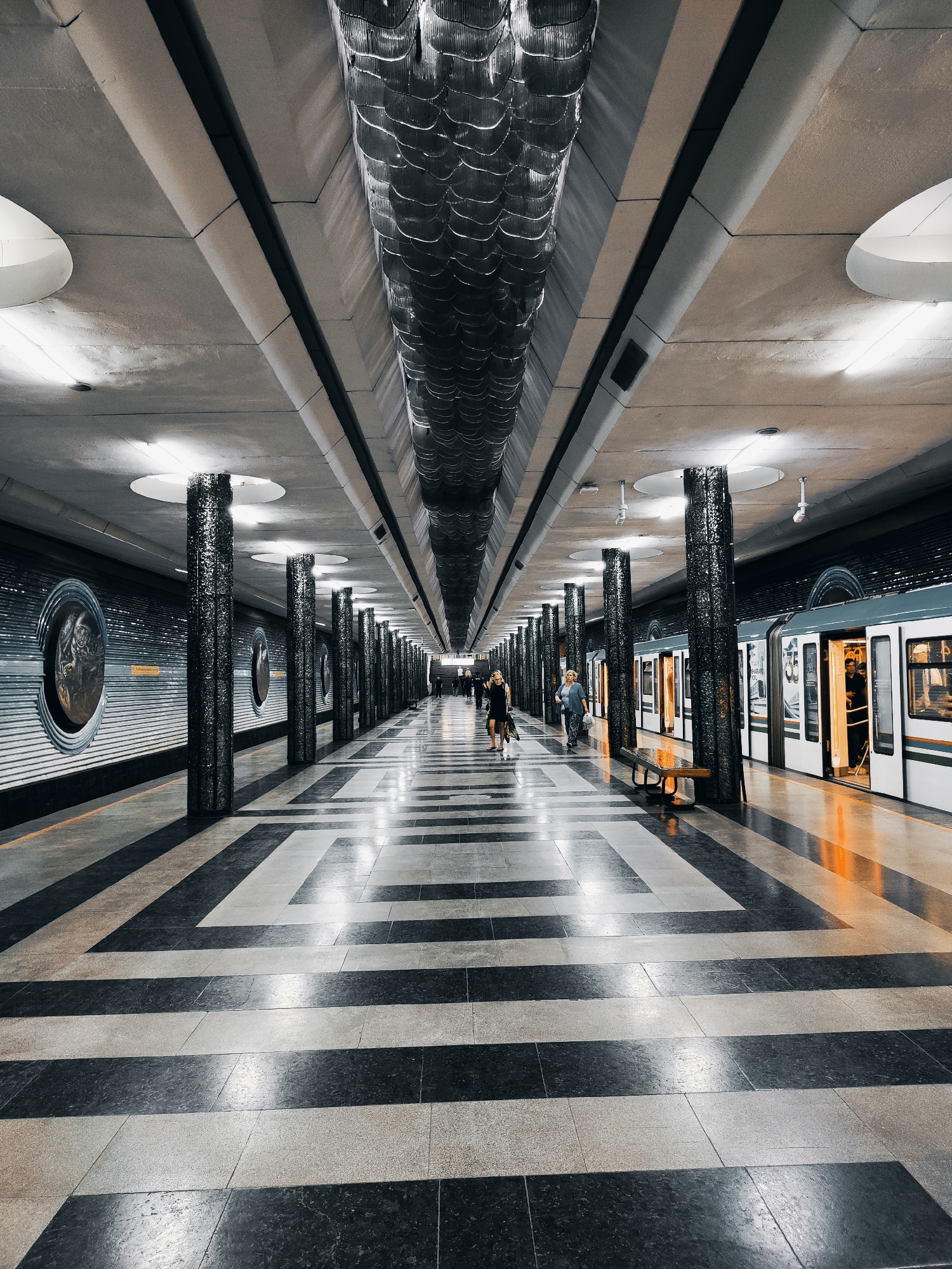 The image shows a wide, empty metro station platform in Tashkent, Uzbekistan. The station features a long, narrow layout with a symmetrical design. The floor is covered in a striking black and white geometric tile pattern. On either side of the platform are rows of tall, dark, textured pillars that support a high, arched ceiling. The ceiling is adorned with decorative, wave-like metallic structures.
On the left side of the platform, a wall is decorated with large, circular relief sculptures and horizontal paneling. On the right side, a train is stopped at the platform with its doors open, revealing passengers inside. A few people are visible on the platform: two women are walking in the foreground, and a couple more individuals are seen further down the platform near the train. The lighting is a mix of overhead illumination and the warm glow from the train's interior, casting reflections on the polished floor. The overall impression is one of grand, somewhat austere, modernist architecture.