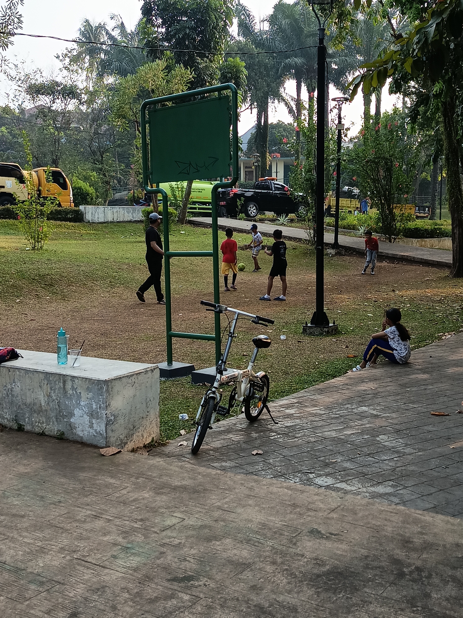 In a park in Depok, Indonesia, several individuals are engaged in various activities. A young boy in a red shirt and yellow shorts kicks a soccer ball, while two other boys, one in black and the other in white, stand nearby. Further in the background, a man in a black shirt and pants walks across the grassy field. A girl in a white shirt and blue pants sits on the paved walkway, observing the scene. A folding bicycle is parked on the grass near the walkway, and a blue water bottle rests on a concrete bench. In the background, construction vehicles and a black pickup truck are visible. The environment is a green park with trees and a paved walkway.
