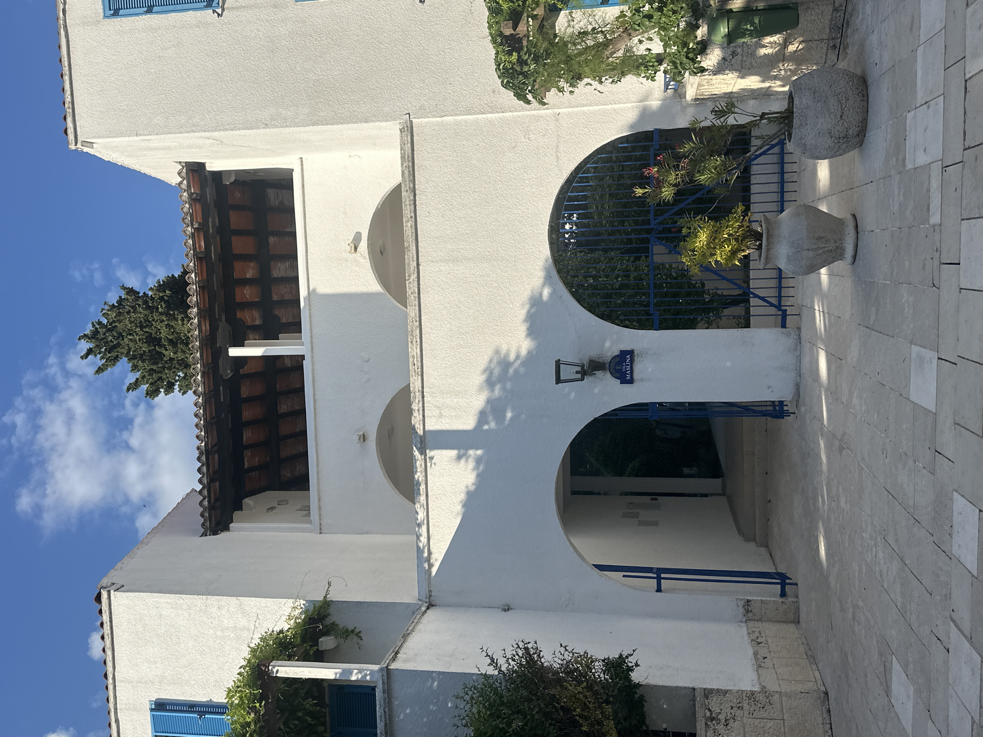 The image shows the entrance to Vila Maslina in Budva, Montenegro. The building is white with blue shutters and a red-tiled roof. There are two arched entrances, one leading to a walkway and the other to a courtyard with a blue gate. Two potted plants are placed in front of the courtyard entrance. The ground is paved with stone tiles. The sky is blue with some clouds, and sunlight casts shadows on the building and pavement.