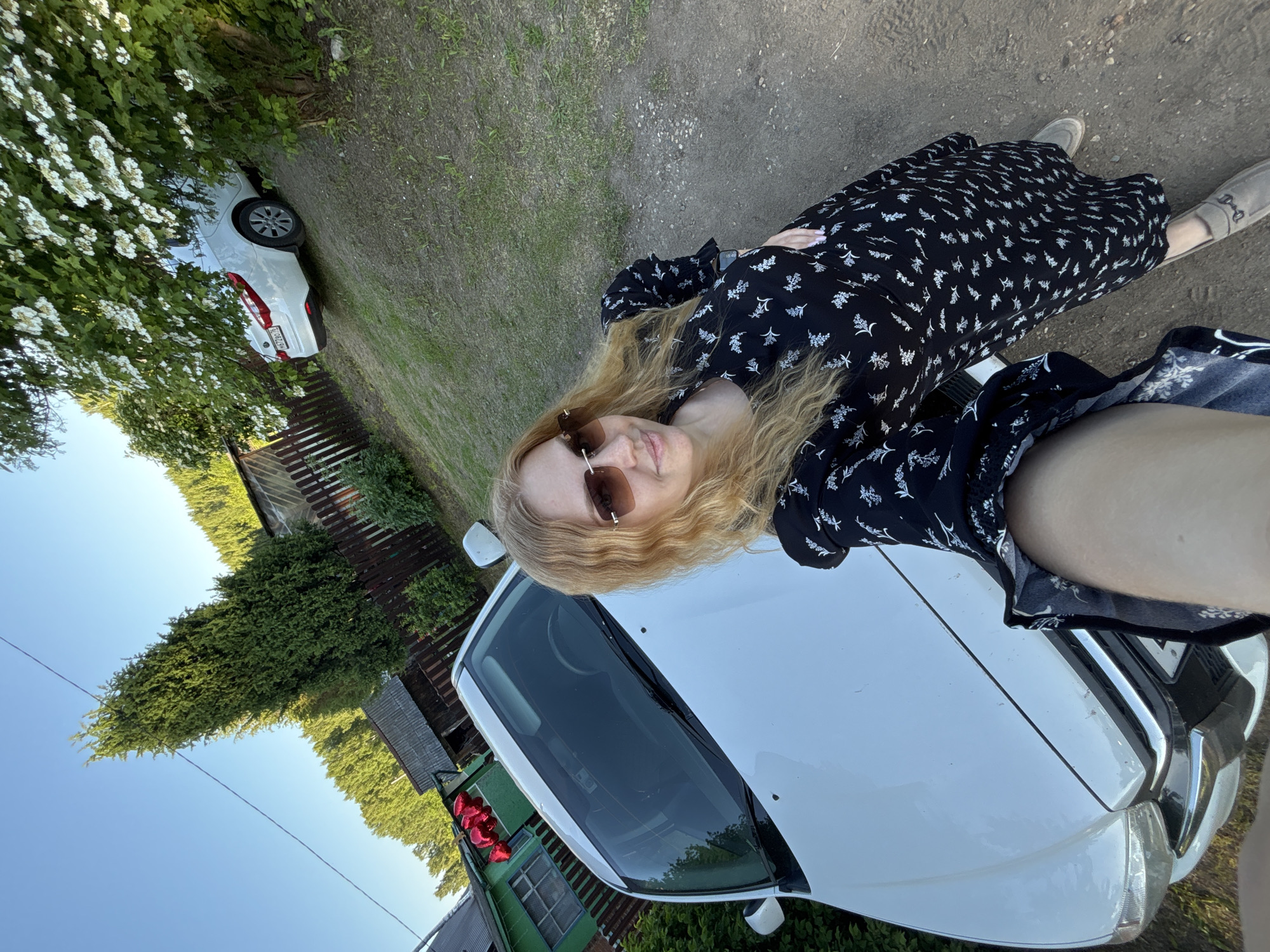 A woman is captured in a selfie in Lepilovo, Russia. She has long, wavy blonde hair and wears sunglasses with brown lenses and a gold frame. Her attire consists of a black dress patterned with white floral or leaf motifs. Her fingernails are painted a light color, and she wears light brown loafers. The woman holds the camera at an elevated angle, showing her arm partially in the lower left, and presents a slight smile.
In the foreground, a white car, possibly a sedan or small SUV, is parked, facing the camera. Behind this car and to the left, a small green wooden building with brown trim is visible, in front of which a cluster of red heart-shaped balloons is attached. Further back, a rustic wooden structure with a corrugated metal roof stands near a dark brown wooden fence. Tall, narrow evergreen trees, possibly cypress, line the property and a distant hillside.
In the background, on a grassy incline partially obscured by a flowering bush with white blossoms, another white sedan is parked. Its rear license plate, "H472BO76", is visible. The sky in the upper left is clear blue, and a thin black wire stretches diagonally across it. The ground is composed of a dirt path mixed with patches of grass. The scene is illuminated by natural daylight.