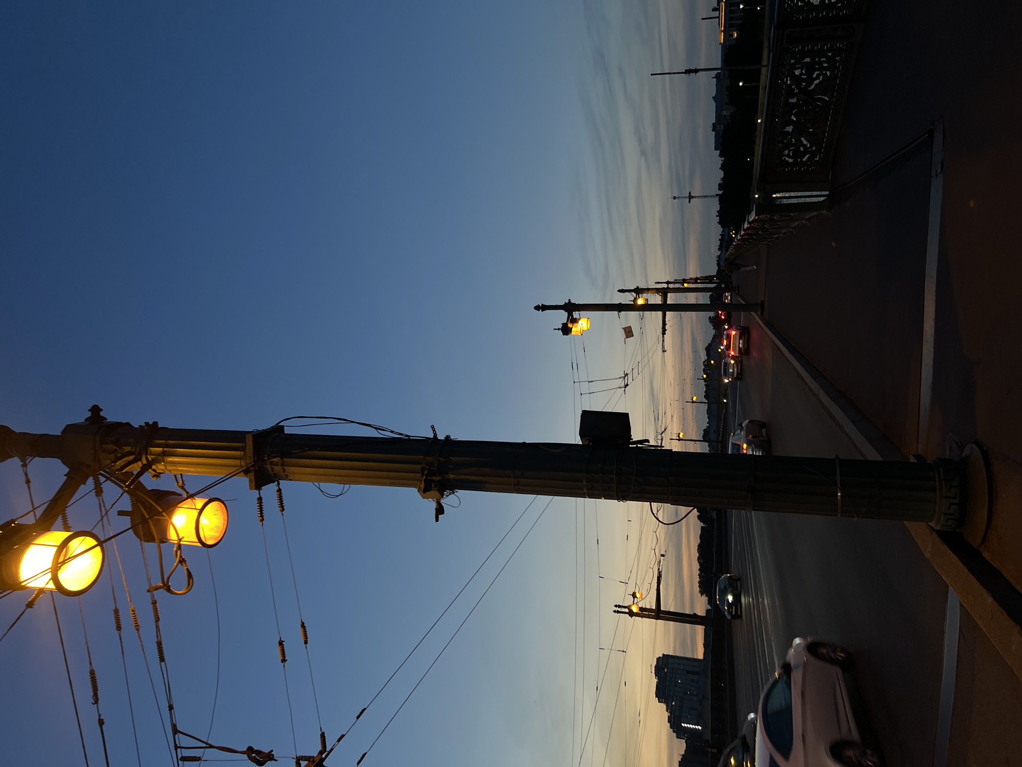 The image shows a view of a bridge at dusk in an unspecified city in Russia. In the foreground, a large lamppost with three glowing yellow lights stands prominently on the right side. The bridge has a pedestrian walkway on the right with an ornate railing. On the left, a roadway is visible with several cars driving by, their headlights and taillights illuminated. The sky is a deep blue, transitioning to lighter shades near the horizon where a faint sunset glow can be seen. In the distance, buildings and more lampposts are silhouetted against the twilight sky. Electrical wires are strung across the scene, characteristic of urban transportation systems. The overall atmosphere is one of a city at the end of the day, with artificial lights beginning to dominate the fading natural light.
