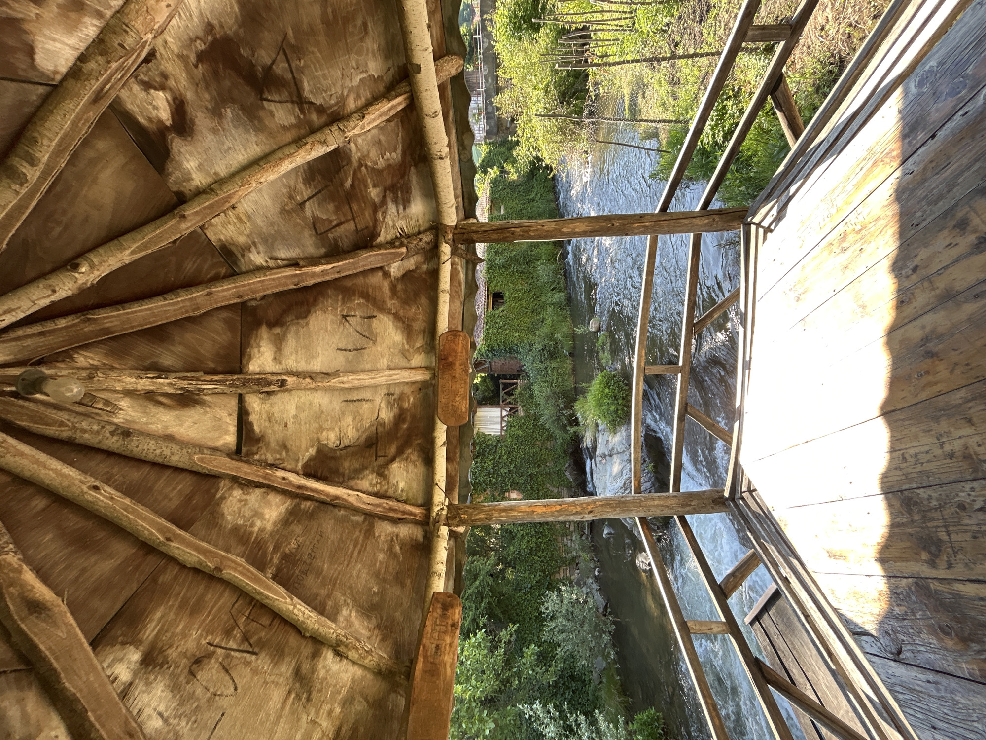 A rustic wooden gazebo overlooks a flowing river. The gazebo's roof, constructed from rough-hewn wood panels and branches, has some markings scratched into its surface. A wooden railing encloses the open-air platform, offering a view of the river and its lush green surroundings. On the far bank, buildings covered in ivy are visible, along with dense vegetation and trees. Sunlight streams through gaps in the gazebo's roof, casting distinct patterns on the wooden floor. The scene appears to be set in a natural, tranquil environment.