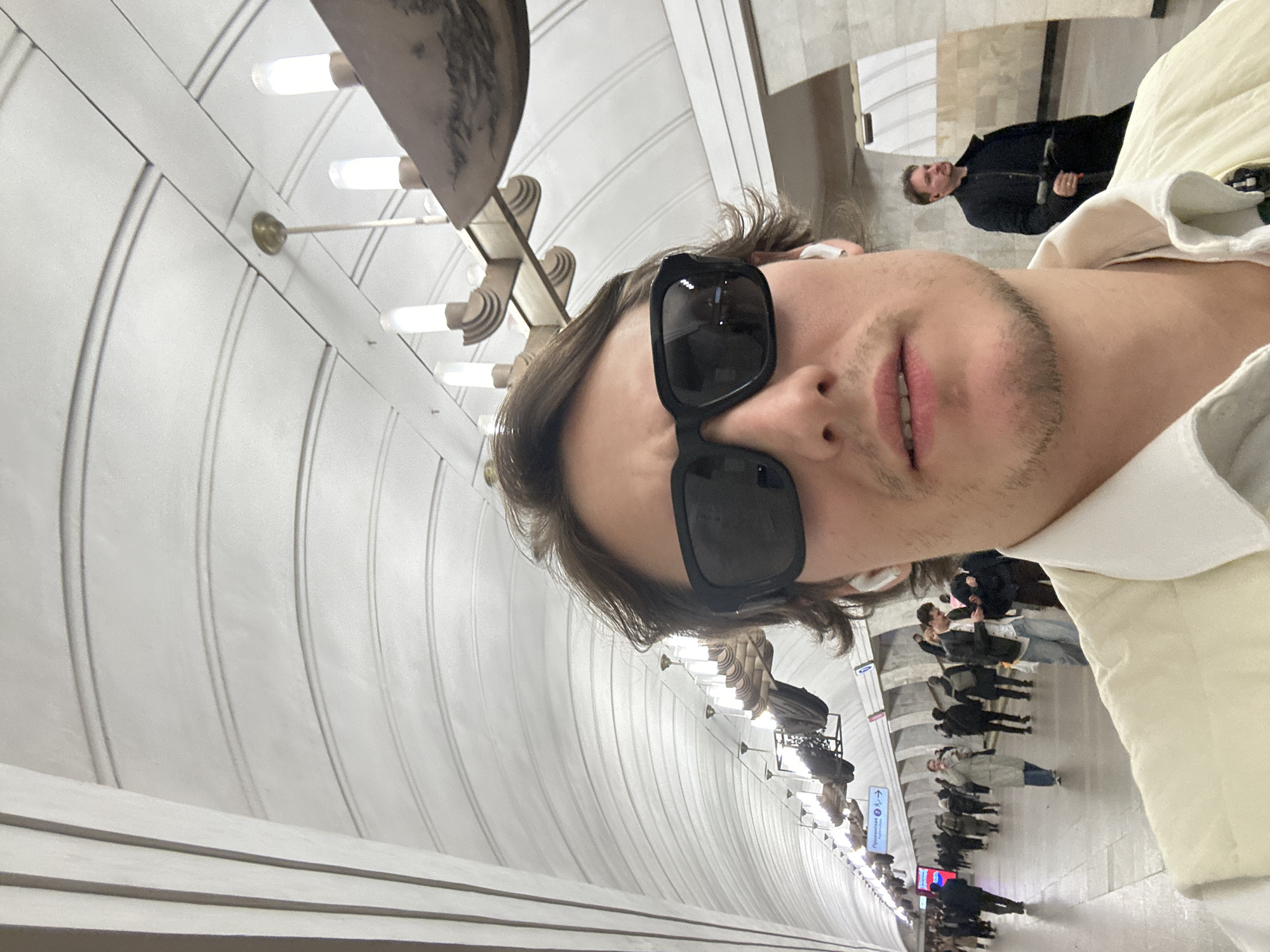 The image depicts a person wearing sunglasses, potentially taking a selfie. The background reveals an interior view of the Moscow Metro in Russia. Specifically, the curved, white tiled walls with decorative lighting fixtures indicate that this is likely the Arbatskaya station on the Arbatsko-Pokrovskaya Line. Other people are present in the background of the underground subway station. The floor appears to be a light-colored stone or tile. Signage is visible in the background with arrows in what appears to be Russian.