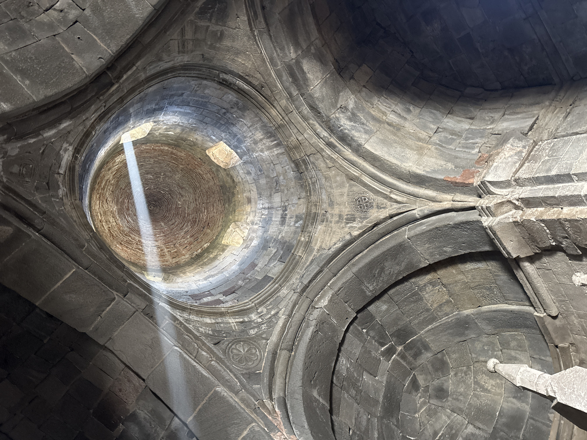 The image depicts the interior dome of the Holy Trinity Church, also known as Gergeti Trinity Church, in Stepantsminda, Georgia. The dome is constructed of dark grey stone, with a circular opening at the top allowing a beam of sunlight to penetrate. The interior surface of the dome is made of reddish bricks in a spiral pattern. The light highlights the texture and stonework of the dome.