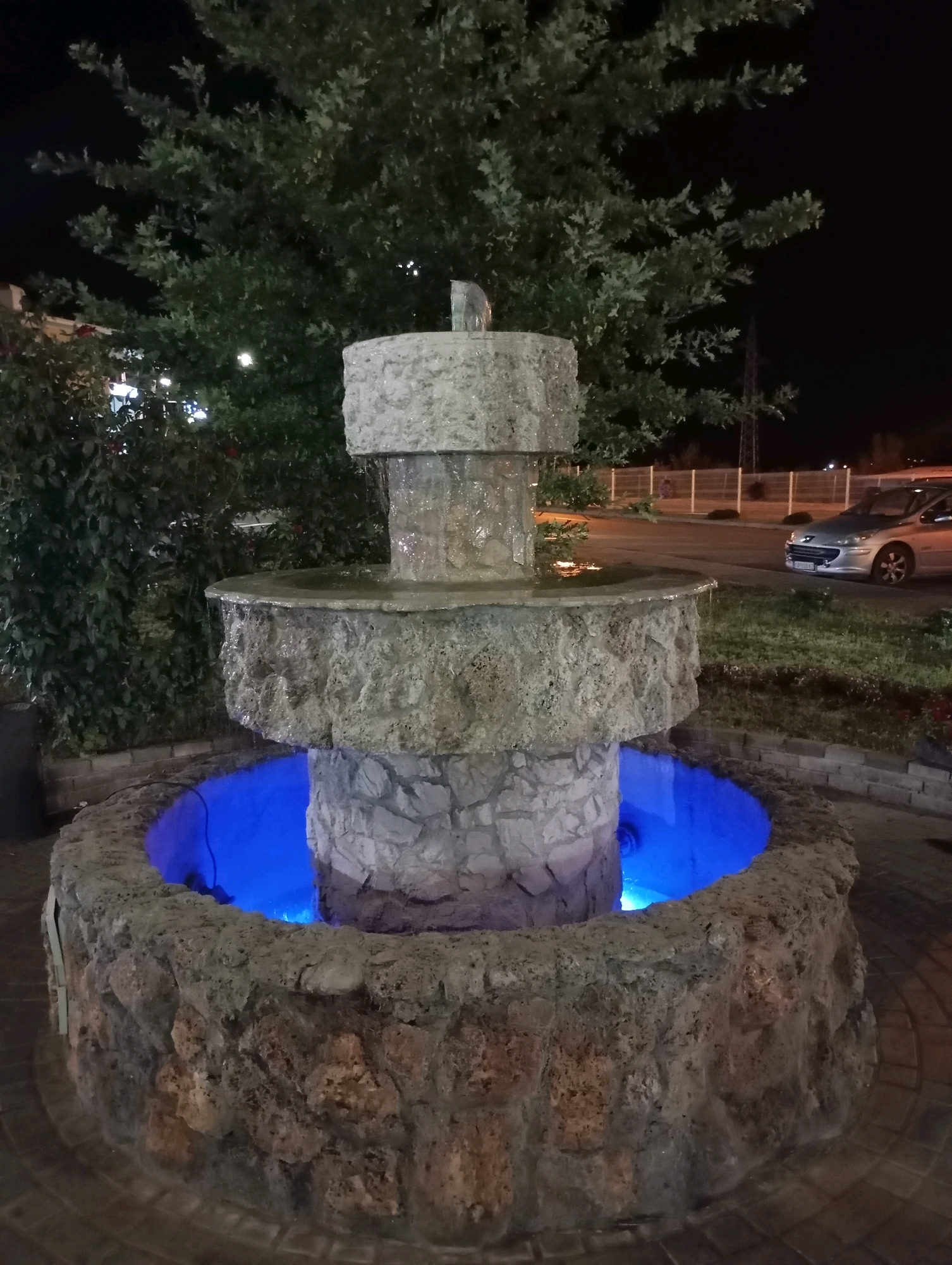 The image shows a multi-tiered stone fountain at night. The fountain has three levels, with water cascading down from the top. The basin of the fountain is illuminated with a bright blue light. In the background, there are trees, a fence, and a silver car parked on the side of the road. The scene appears to be in an outdoor public space, possibly a park or a plaza. The location is identified as Topola Municipality, Serbia.