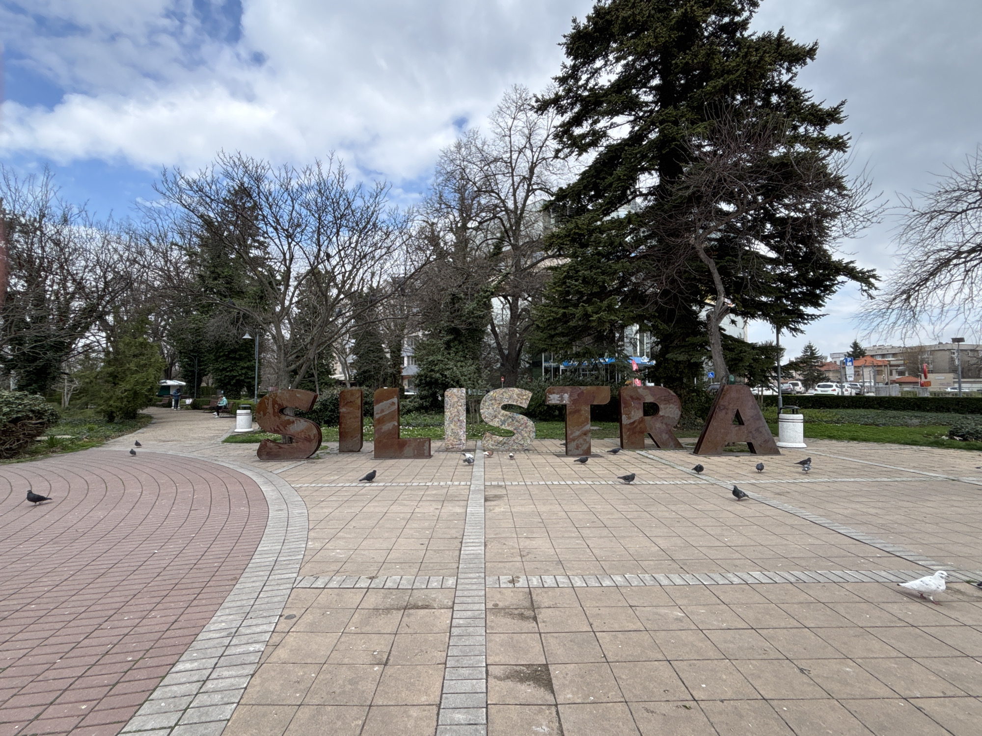 The image shows a public square in Silistra, Bulgaria. Large, weathered, brown metal letters spelling "SILISTRA" are prominently displayed in the center of the square. Pigeons are scattered around the letters and across the paved square. The paving consists of light gray and red bricks. Trees and some greenery surround the open space, along with some buildings in the background. The sky is partly cloudy. In the back ground, there is a white trash can and there are cars parked on the street.