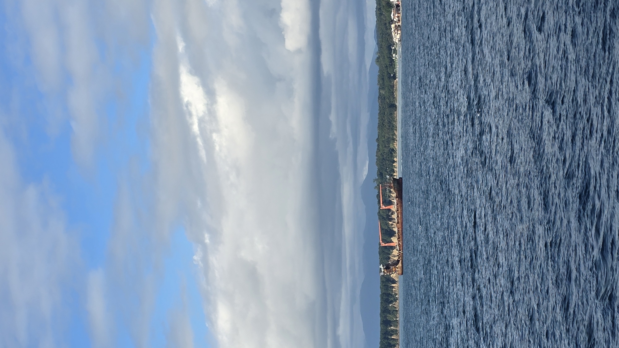 A cargo ship, appearing to be a sand carrier due to its open hull and visible material, is positioned on the water. The ship has a rust-colored hull and a prominent orange crane structure. In the background, a coastline is visible, characterized by a line of trees and hilly terrain extending into the distance. Further along the coastline, small buildings suggest a populated area. The sky is partly cloudy with a mix of blue sky and white and gray clouds. The water is choppy and dark blue. The scene is likely located in the vicinity of Nakhodka, Russia, given the characteristic coastline and maritime activity. No people are discernible in the image.