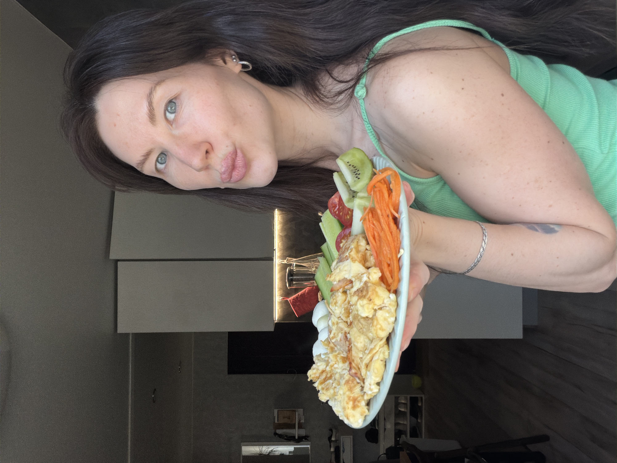 A woman with long, dark hair and blue eyes poses indoors, holding a plate of food. She wears a light green tank top and a silver bracelet on her left wrist. The plate contains scrambled eggs, kiwi slices, cucumbers, tomatoes, and shredded carrots. The background shows a modern kitchen with gray cabinets and hardwood floors. The city is Perm, Russia.
