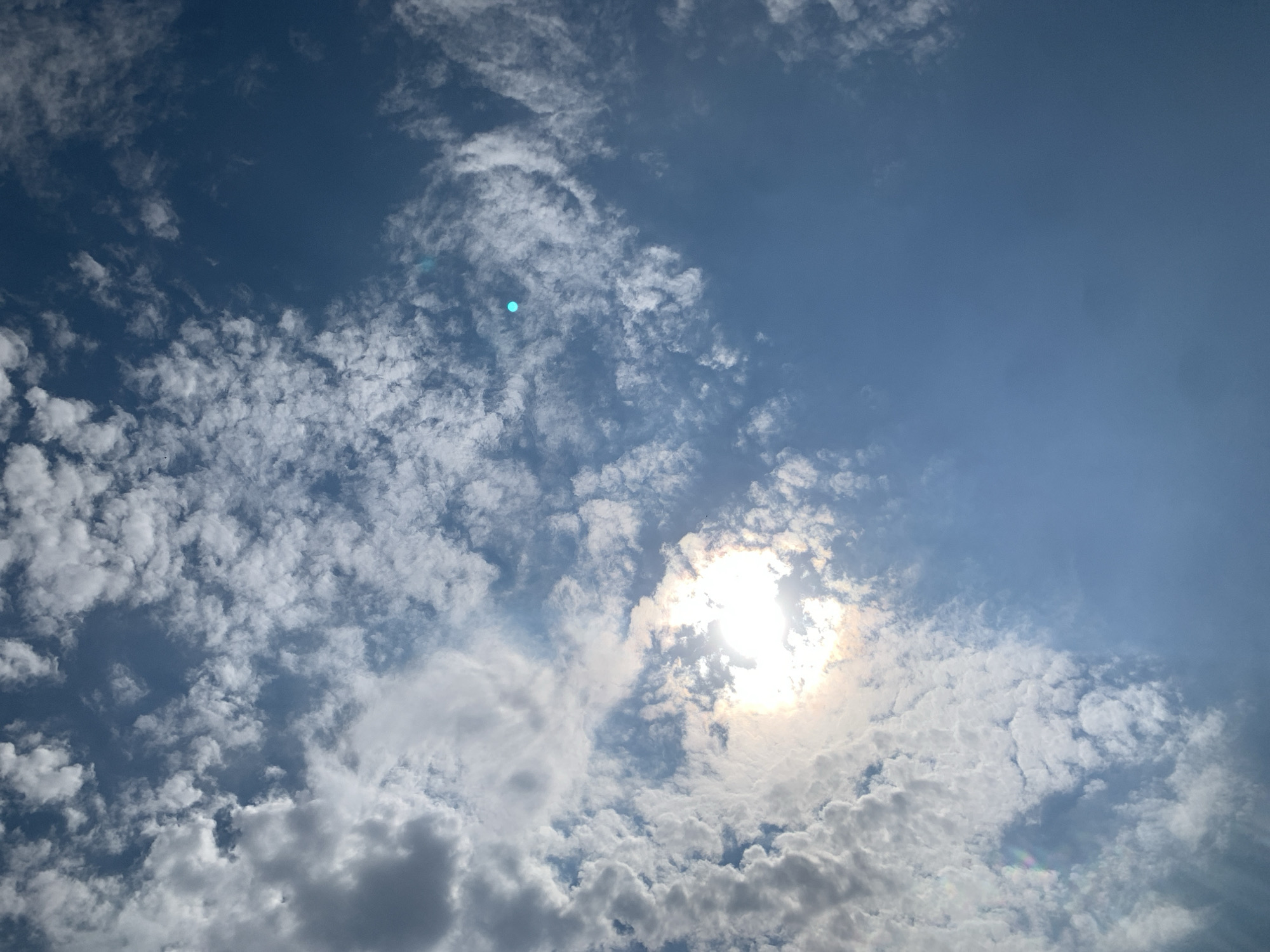 The image captures a bright, sunny sky with scattered clouds. The sun is visible through a break in the clouds, creating a strong glare. The sky is a vibrant blue. There are no people or man-made objects visible in the frame. The scene is entirely natural, focused on the sky and clouds. There is no information within the image to suggest it is located in Khazar Raion, Azerbaijan; the description is purely based on the visual content of the photograph.