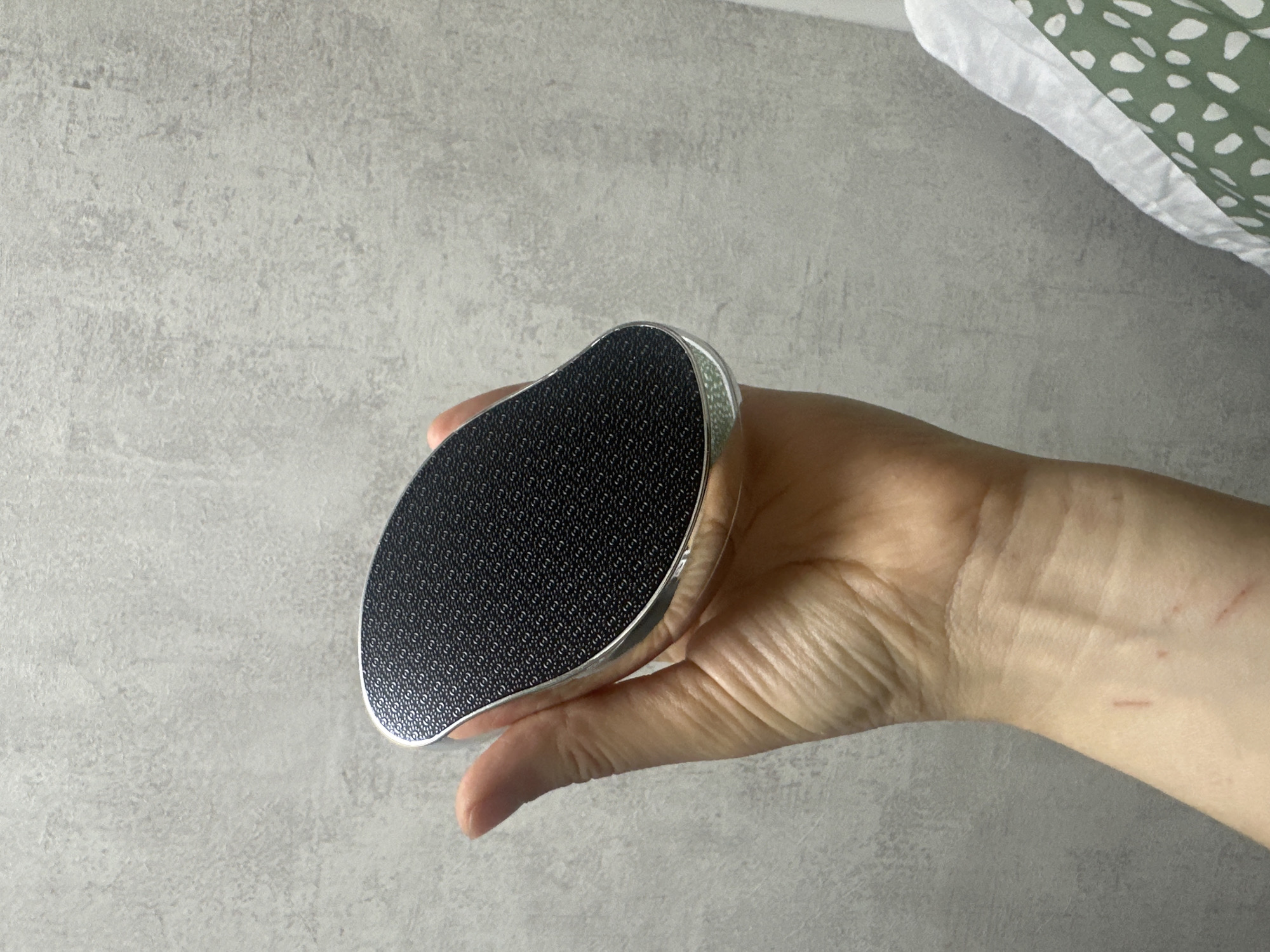 This media file shows a close-up of a hand holding a foot file. The foot file has a black textured surface with a repeating pattern and a silver-colored border. The hand appears to be a person's left hand, with visible lines on the palm. The background is a textured gray surface, possibly a wall or countertop. In the upper right corner of the image, a white and green patterned fabric is visible.
The activity taking place is the presentation of the foot file, held by the hand. There are no clear indicators of the time of day or weather. The location context is given as Moscow, Russia. No text is visible in the image. The overall impression is that of a product being displayed.