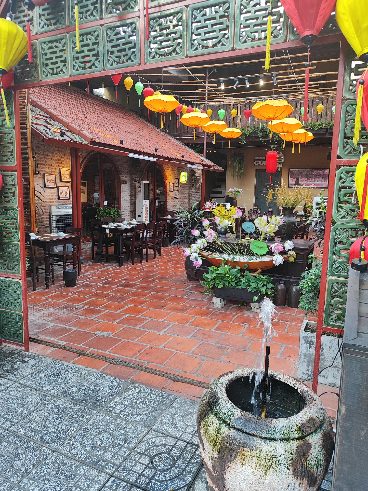 The image depicts the outdoor seating area of a restaurant in Da Nang, Vietnam. The space is paved with reddish-brown square tiles, and there is a smaller section of grey patterned tiles in the foreground. The area is decorated with numerous colorful lanterns, primarily yellow and red, hanging from bamboo poles. A traditional Vietnamese architectural style is evident in the green lattice work and the red-tiled roof of the building.
Several wooden tables and chairs are arranged on the tiled floor, set up for dining. Some tables have white bowls and plates. In the center of the seating area, a large planter holds an arrangement of artificial lotus flowers and greenery. A small fountain is active in a large, weathered clay pot in the foreground, with water spraying upwards. The building walls are made of brick. A large air conditioning unit is visible on the left side of the building. The overall atmosphere suggests a welcoming and visually appealing dining environment with a distinct cultural influence. There are no people visible in the image.