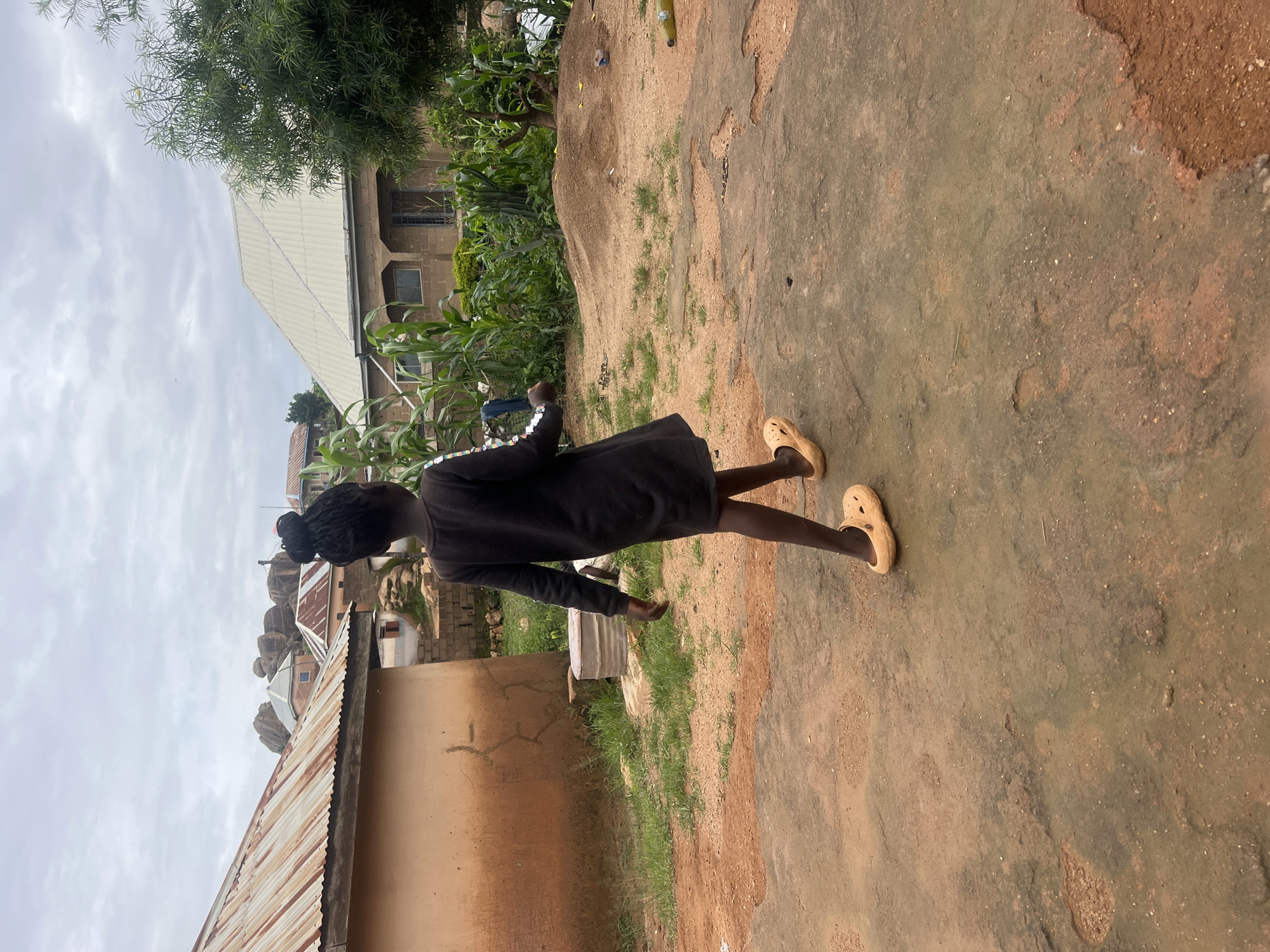 A young girl, facing away from the camera, walks across a dusty, rocky ground in front of a building with a corrugated iron roof. She is wearing a dark, long-sleeved dress and light-colored clogs. In the background, there are more buildings, some with similar corrugated roofs, and a lush green area with corn stalks and trees. The sky is overcast. The scene appears to be in a residential area in Jos, Nigeria.