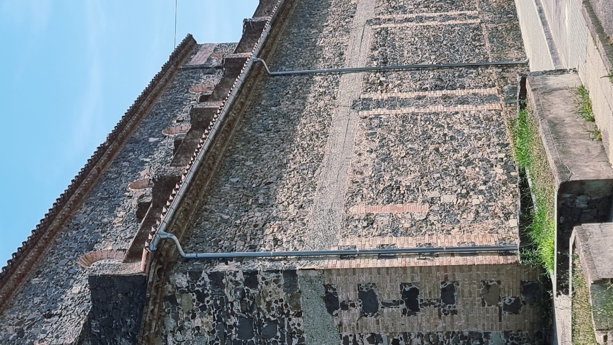 This image depicts the exterior of a building constructed with a mix of stone and brick, likely in Castellfollit de la Roca, Spain. The building has a steep, tiled roofline visible at the top left. The walls are textured with rough-hewn stones and interspersed with horizontal bands of brickwork, some of which appear to be part of an older structure or decorative element.
A metal gutter system runs along the edge of the building, with a pipe extending horizontally across the wall. This suggests it's a functional aspect of the building's design, likely for water drainage. In the lower right portion of the image, a concrete structure with channels is visible, overgrown with green grass. This might be part of a drainage system or terracing leading away from the building.
The sky above is a clear, light blue, indicating fair weather, likely during the daytime. The lighting suggests a sunny day, casting subtle shadows that highlight the textures of the building materials. There are no people or animals visible in the frame, and no text is present. The scene appears to be a detail of the building's facade and its immediate surroundings, focusing on architectural features and materials. The overall impression is one of aged construction and a natural integration with its environment, given the greenery.