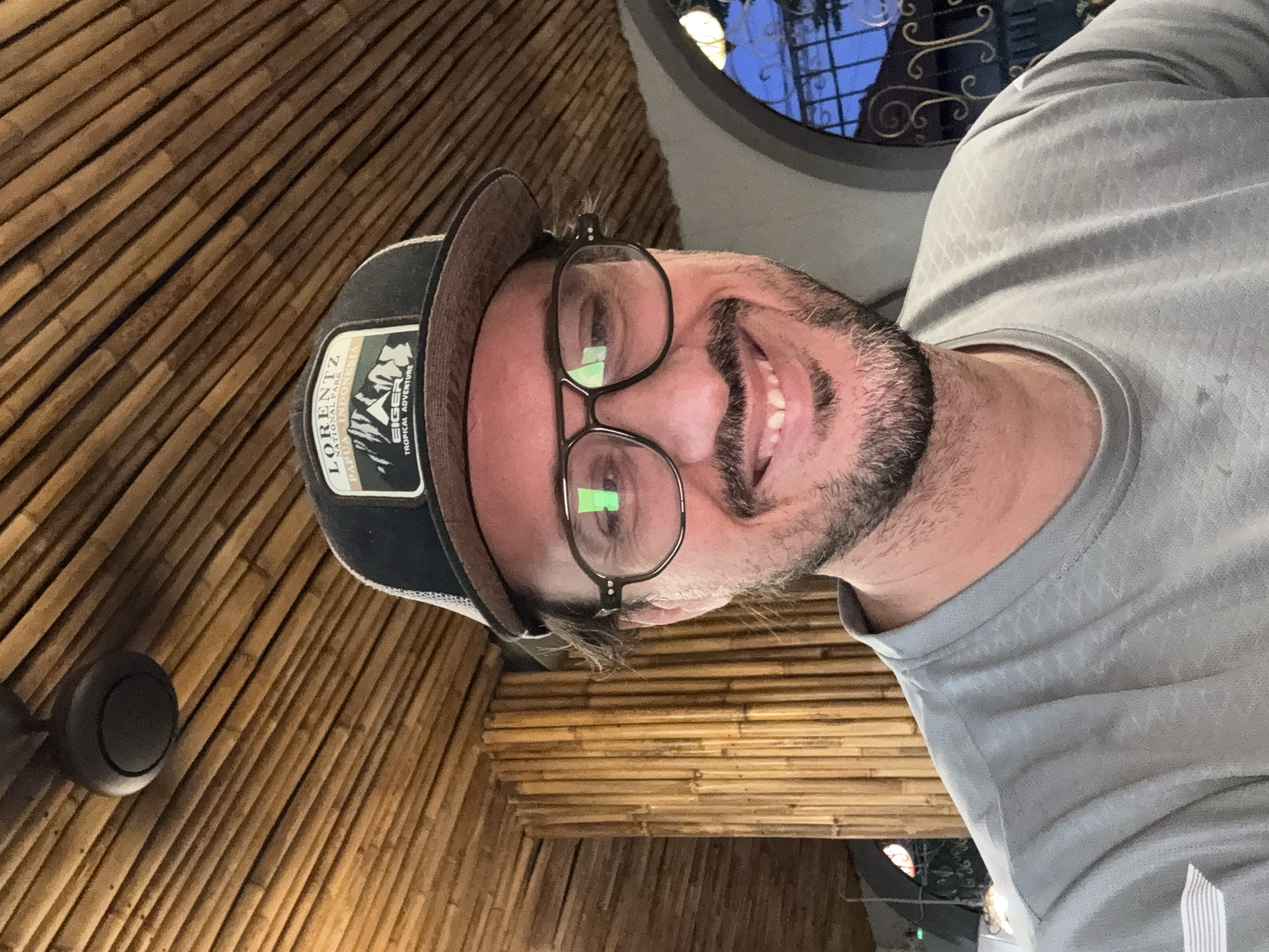 A man with glasses and a baseball cap is smiling at the camera. The cap has a patch on the front that reads "Lorentz National Park Papua Indonesia" and "EIGER Tropical Adventure". He is wearing a gray t-shirt. The background is comprised of bamboo. The setting appears to be indoors. Based on the textual details on the cap, the location is potentially within the vicinity of Lorentz National Park in Papua, Indonesia, although Kuta Selatan is in Bali.