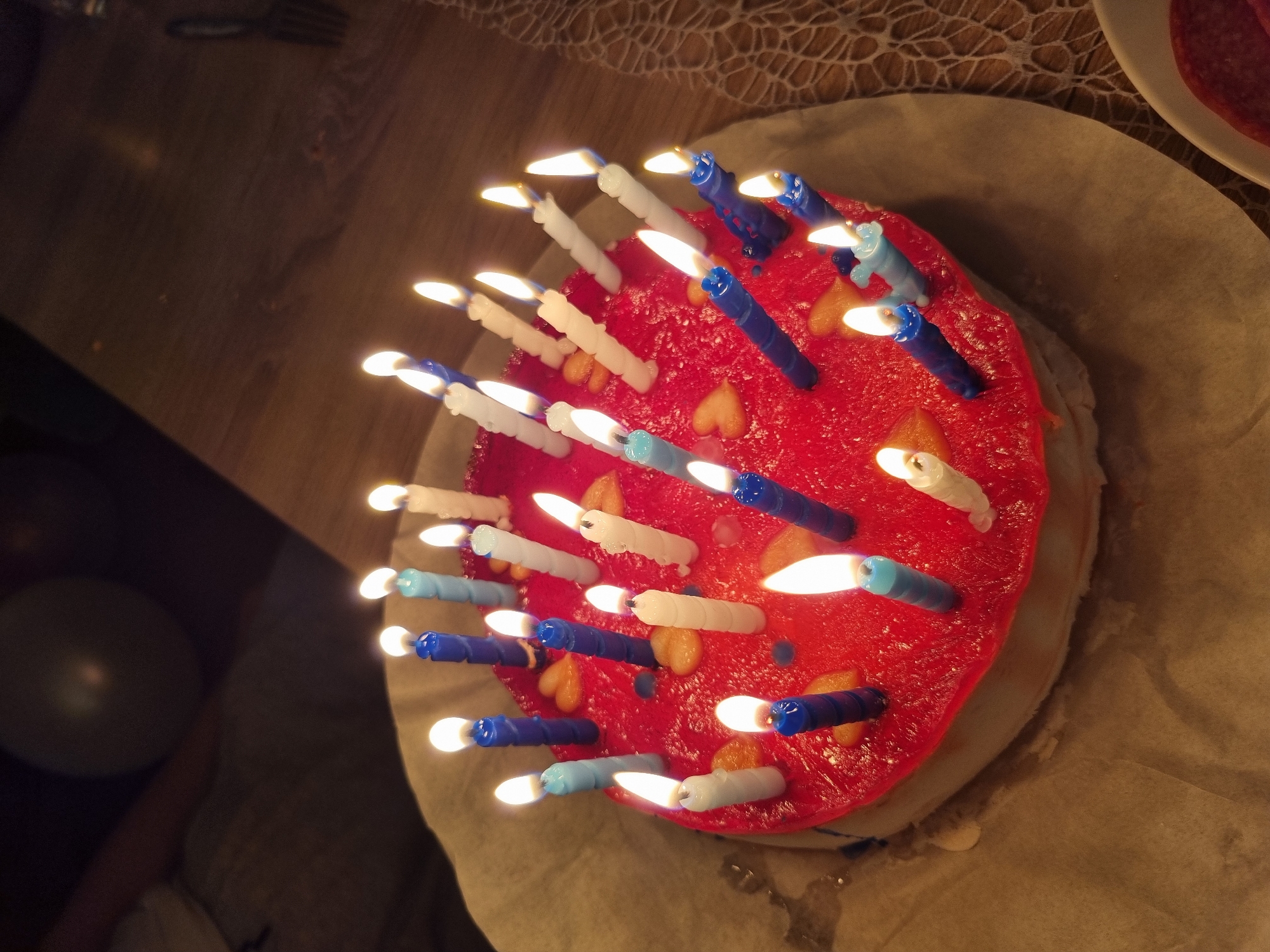 A high-angle shot shows a birthday cake with many lit candles, positioned on parchment paper. The cake has a vibrant red frosting. Scattered around the cake are small heart-shaped decorations, alongside what appears to be small pieces of candied fruit. The candles are a mix of white, blue, and light blue colors, all burning brightly. In the upper right corner, a plate with slices of salami is partially visible. In the background, on the left side, there is a partially visible, round, gray-colored balloon. In the upper left corner, a part of a fork is visible on a dark brown wooden surface. There is no indication of the location being Saarbrücken, Germany. There are no people visible in the image.