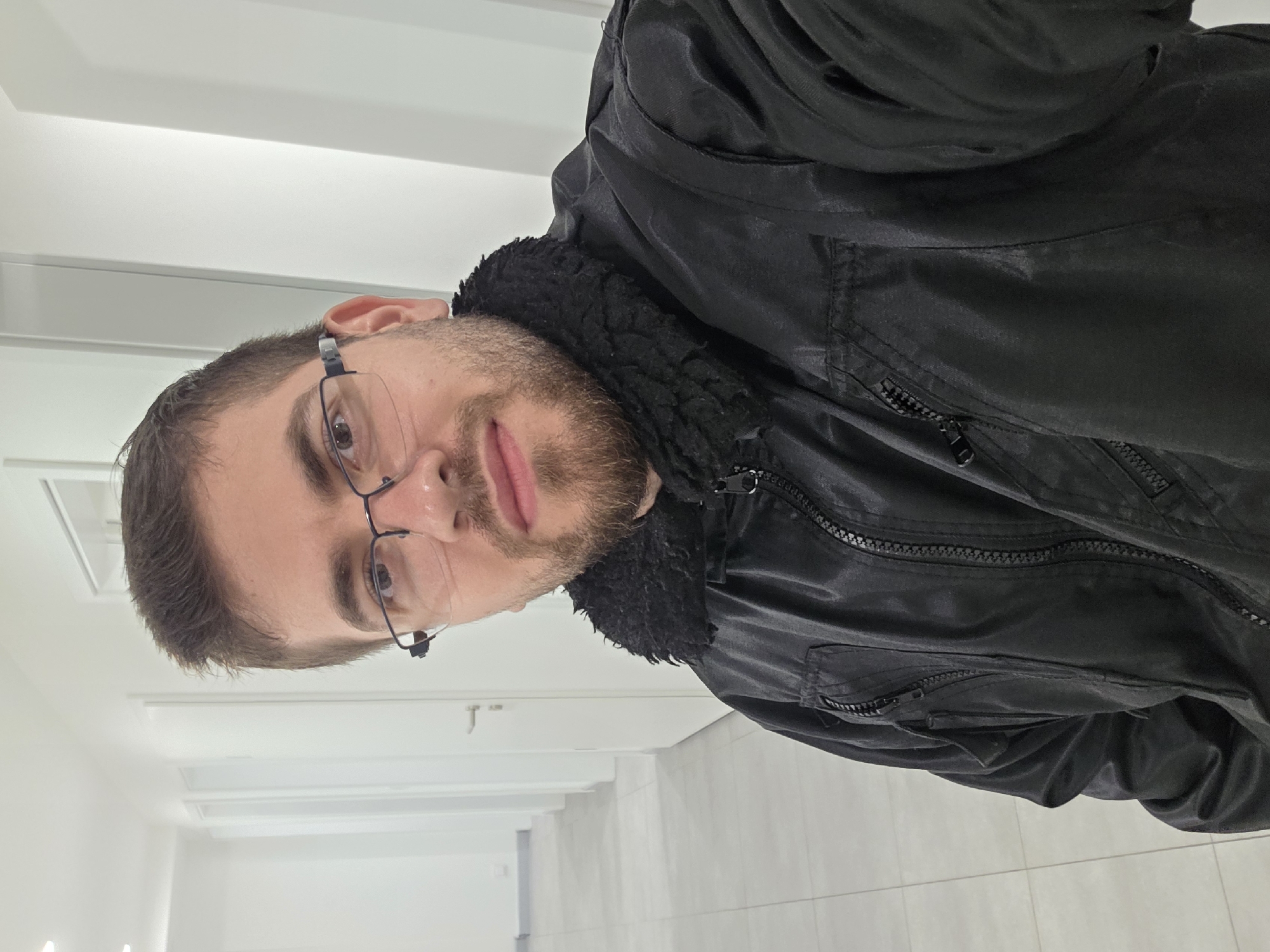 A man with fair skin, a beard, and glasses stands indoors. He is wearing a black jacket with a black, fur-like collar. The background appears to be a hallway with white walls and a tiled floor. The location is not identifiable as being in Plovdiv, Bulgaria from the provided image.