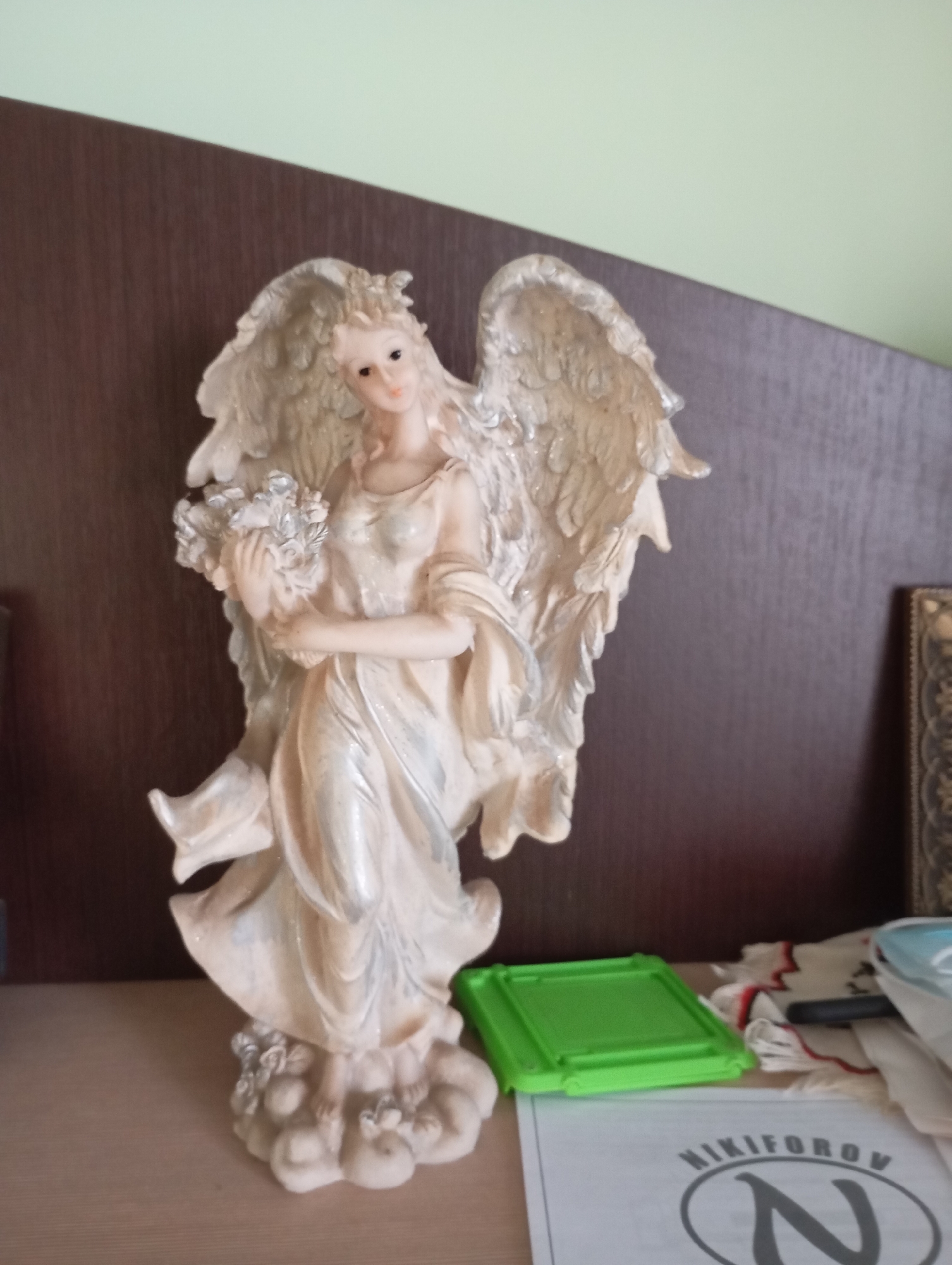 A static shot shows a detailed figurine of a light-skinned angel with feathery wings, a flowing gown, and a floral bouquet. The angel is positioned on the right side of the frame, standing on a cloud-like base. To the right of the figurine, a green rectangular object rests on a surface, with a white cloth featuring red and black accents partially visible behind it. A light-colored sheet with text and a stylized "N" logo is in the foreground. The background consists of a dark wooden panel and a light green wall. The scene appears to be an indoor setting, possibly a shelf or desk, in a residence located in Horishni Plavni, Ukraine.