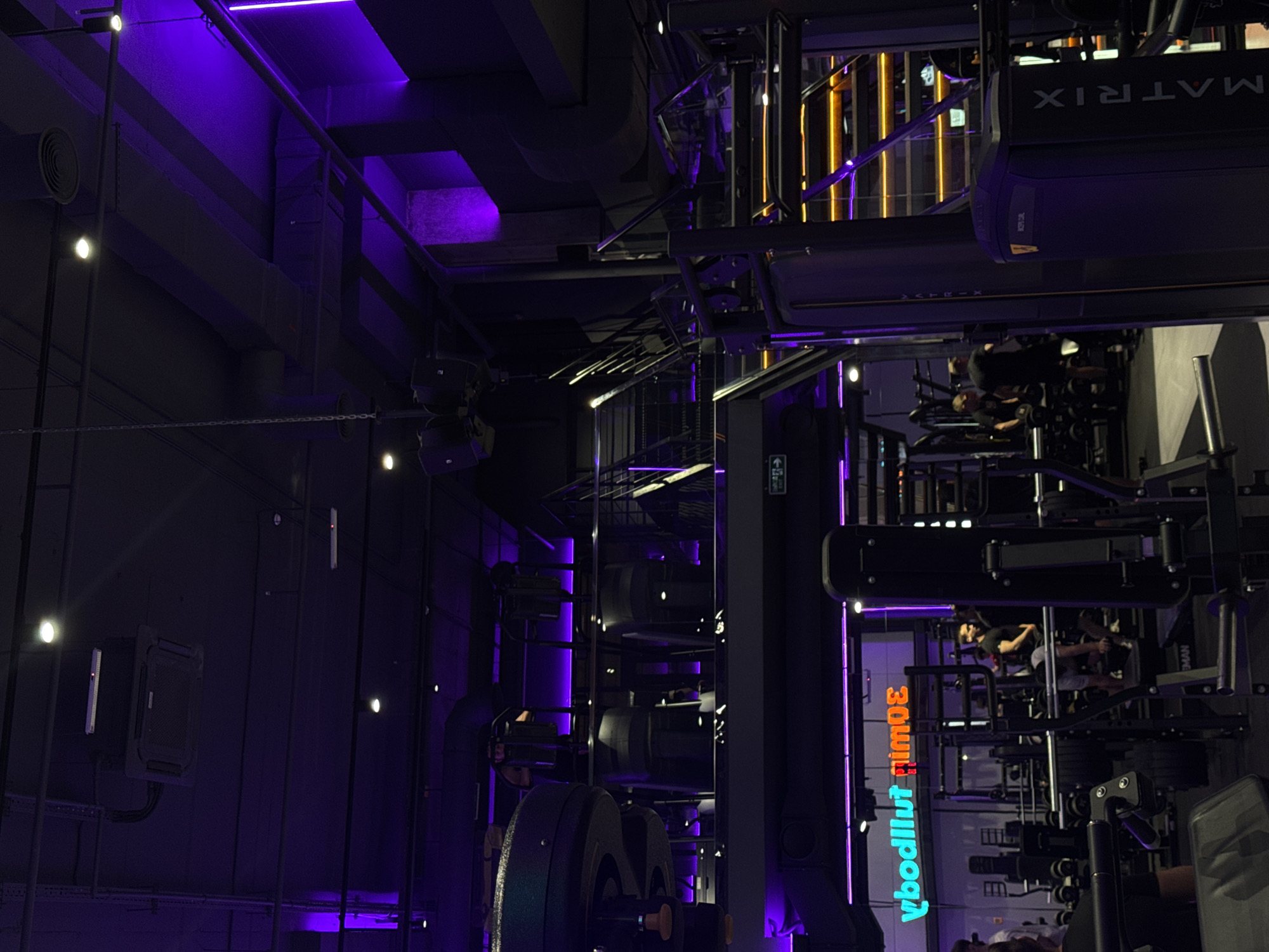 The image depicts the interior of a dimly lit, modern gym, bathed in vibrant purple and orange neon lighting. The gym appears to be multi-level, with a prominent staircase featuring orange illuminated steps. Various weightlifting equipment, including racks, benches, and machines, are visible throughout the space. Several individuals, dressed in athletic wear, are engaged in or observing exercise routines. A neon sign with Cyrillic lettering is mounted on a wall. The overall aesthetic suggests a high-end fitness facility. The location is identified as the "Sport Life" gym in Kazan, Russia.