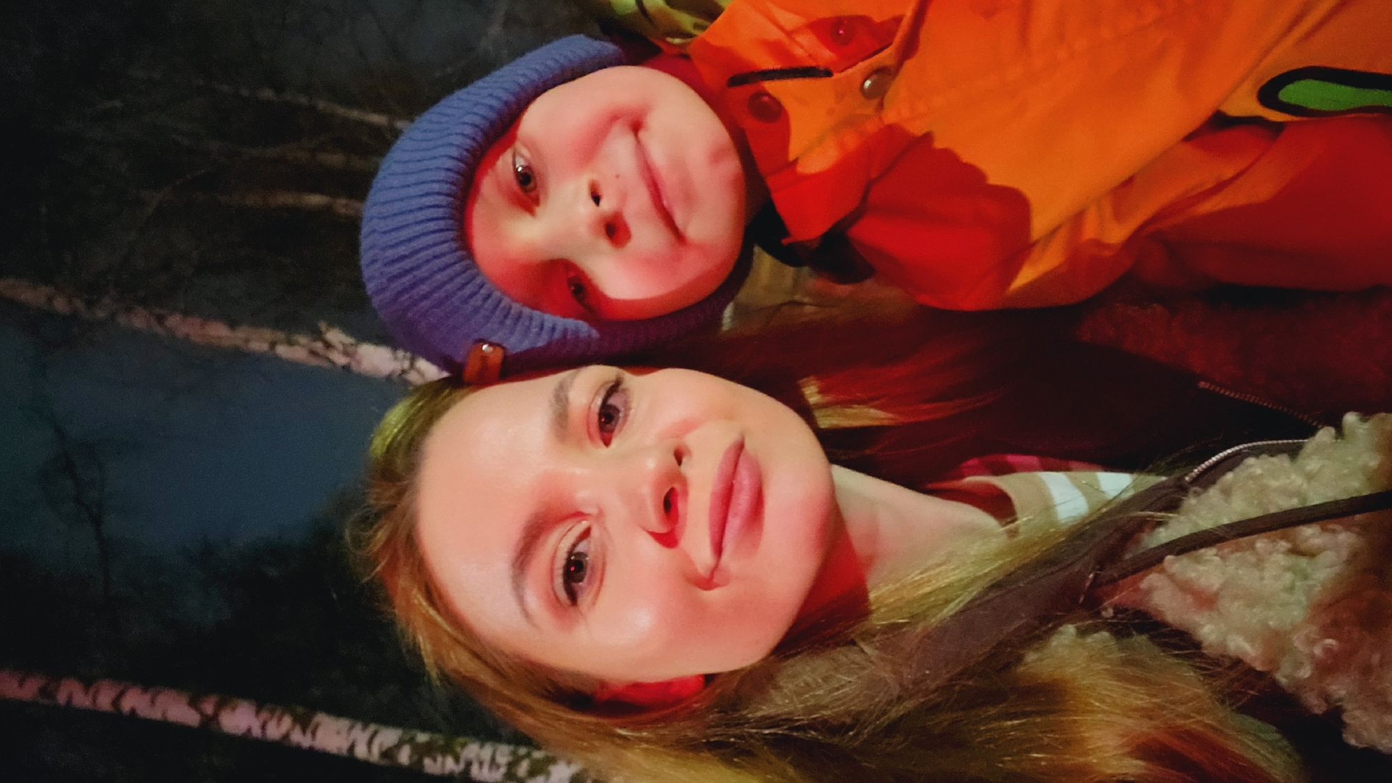 A selfie captures a woman with long blonde hair and a child wearing a blue hat and an orange jacket. They appear to be outside in the evening or at night, possibly in a park or wooded area, given the dark background and faint suggestion of trees. The location is identified as Chaikovsky, Russia.