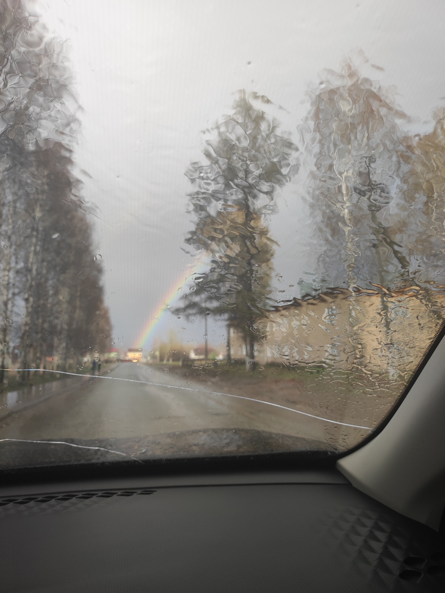 The image depicts a view through a rain-splattered windshield, likely of a car, revealing a street in Votkinsk, Russia. Rain distorts the view, and a rainbow arches over the road in the distance. Bare trees line the sides of the road, and a building is partially visible to the right. The car's dashboard and part of the interior trim are in the foreground. A crack is visible on the bottom part of the windshield. The scene is overcast and wet, suggesting a rainy day.