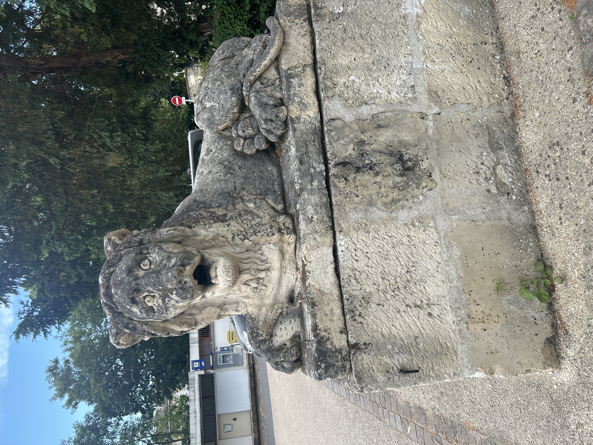A weathered stone sculpture of a lion in a recumbent pose is the central subject, positioned on a large stone pedestal. The lion appears to be roaring, with its mouth open and a somewhat distressed expression. The sculpture is detailed, showing features such as a mane, paws, and a tail curled around its body. The stone itself shows signs of age and weathering, with some discoloration and texture variations.
The sculpture is situated outdoors, with a paved area in the foreground and a background that includes trees and a building. The building in the background appears to be a public facility, possibly a post office or administrative building, with signage visible. A red and white circular "no entry" sign is visible on a post behind the lion, indicating a road or restricted area. The environment suggests an urban or semi-urban setting, possibly a public square or park. The presence of trees indicates a green space within the city.
The overall impression is one of a historical or decorative monument placed in a functional urban environment. The lighting suggests it is daytime, with natural light illuminating the scene. The location is identified as Le Port-Marly, France, adding a specific geographical context to the observation.
