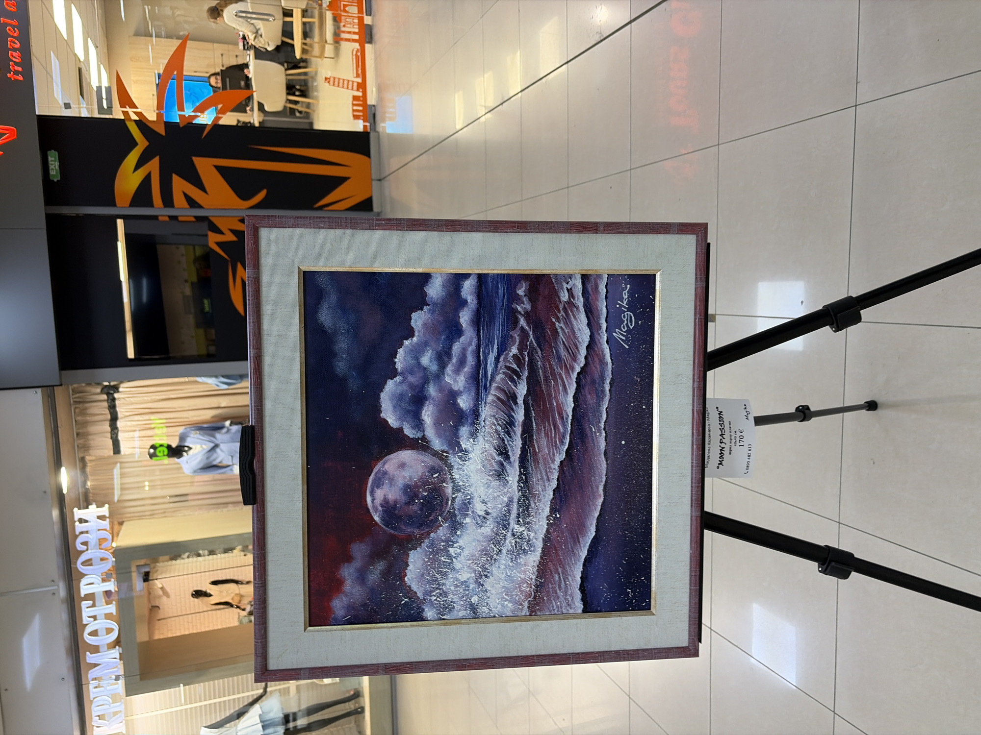 The image shows a framed painting of a moon and wave scene displayed on a black tripod. The painting is signed "Magika". A price tag next to it reads "MOON PASSION" by Magdalena Karayaneva - Madika, acrylic on canvas, 40x40 cm, 170 EUR, with a phone number listed. The painting and stand are placed inside a shopping mall in Plovdiv, Bulgaria. The storefront "КРЕМ ОТ РОЗИ" is visible to the left, and another store with an orange graphic is in the background. A few people are visible further back in the mall. The mall floor consists of light colored tiles. The orientation of the image is such that the right side is higher than the left.