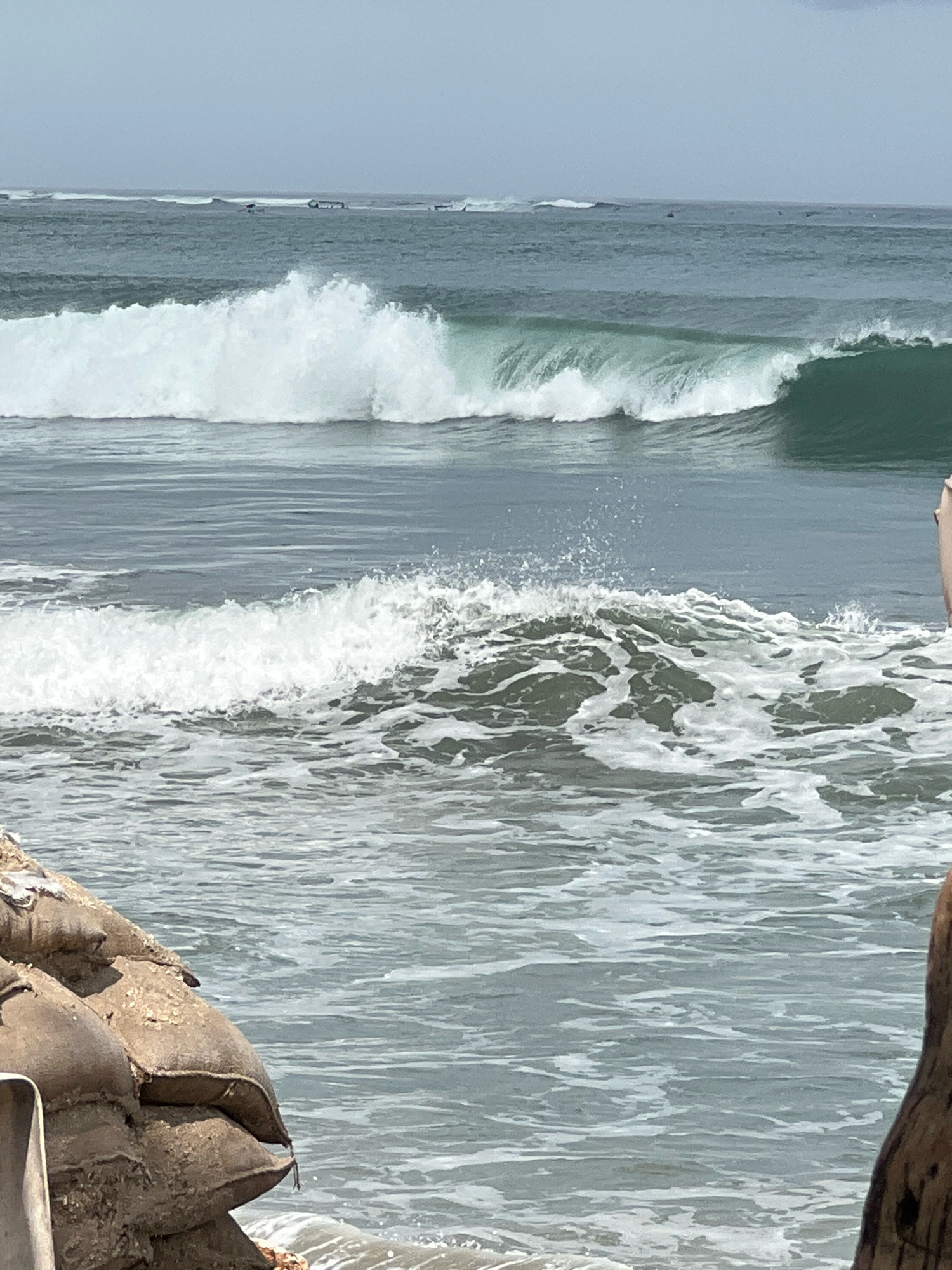 The image depicts a serene beach scene in Kuta, Indonesia, with gentle waves rolling onto the shore. In the foreground, a stack of sandbags serves as a barrier, presumably to protect against erosion or flooding. Beyond the sandbags, the ocean stretches out, with several surfers dotted across the horizon, indicating a popular spot for water sports. The water is a calming shade of turquoise, and the sky is a soft, overcast gray, suggesting a mild, pleasant day for beach activities. The overall atmosphere is one of tranquility and leisure, characteristic of a tropical paradise.