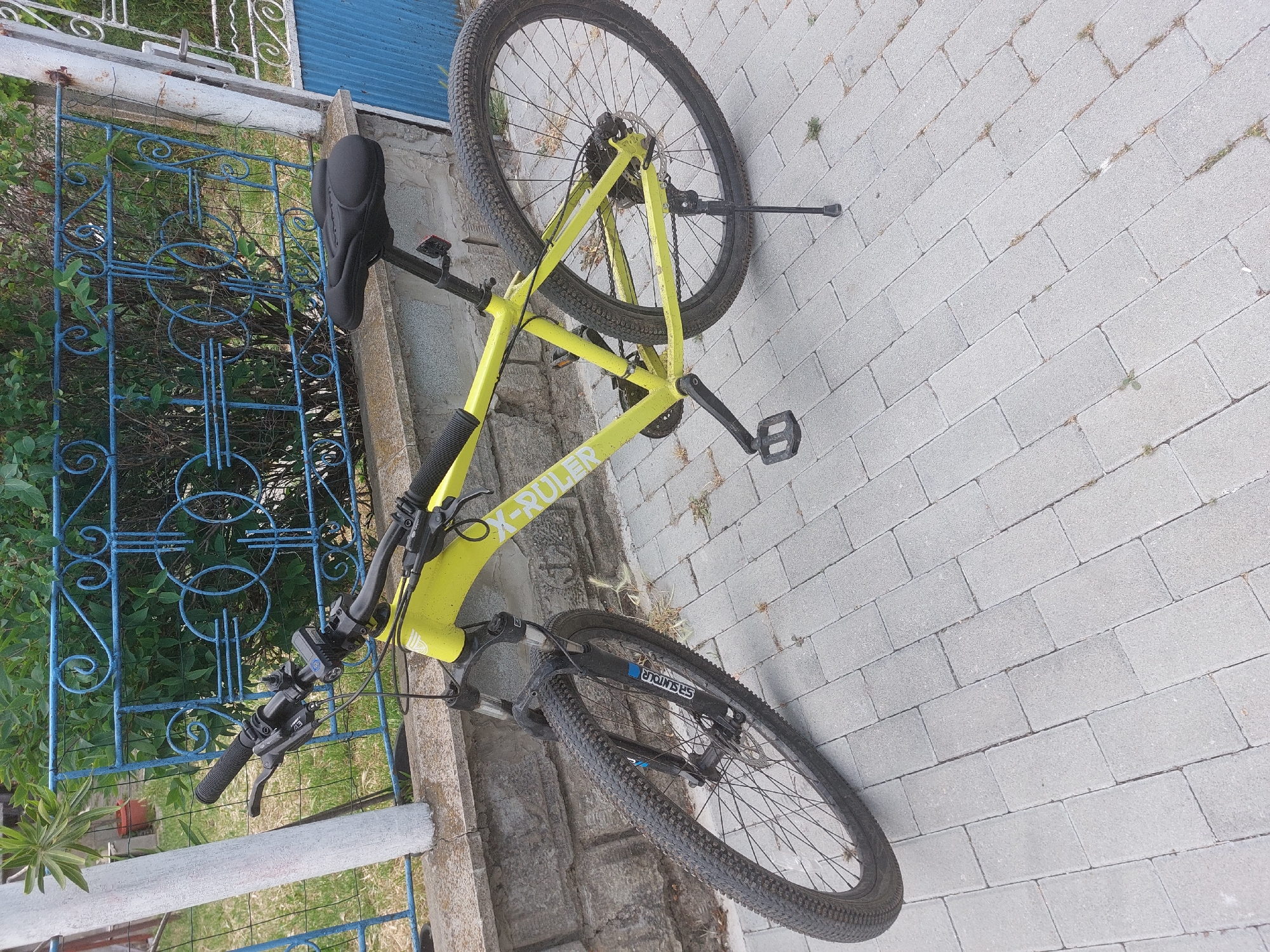 The image shows a yellow X-Ruler mountain bike lying on a grey brick walkway. The bike is positioned between a low concrete wall and a blue metal fence with decorative swirls. Green plants are growing behind the fence. The bike's front tire is oriented towards the bottom right of the image, and its rear tire is near the top. The bike appears to be in Tsarevo, Bulgaria, based on the GPS coordinates.