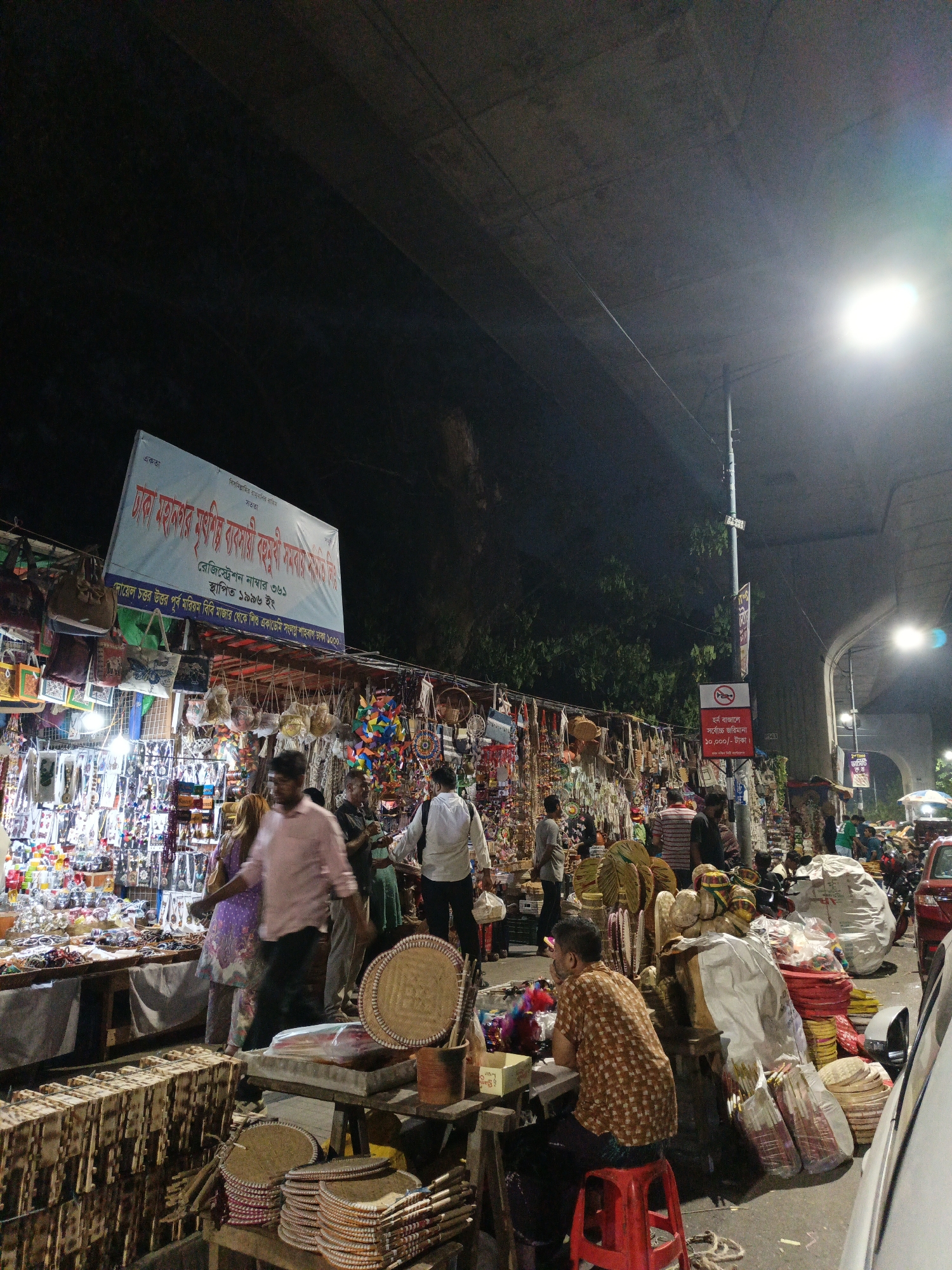 The image shows an outdoor marketplace in Dhaka, Bangladesh at night. Vendors display various goods, including jewelry, bags, and woven crafts, under a bridge. Several people are browsing or walking through the market. A sign reads "Dhaka Mahanagar Murti Bastabi Kamthi Samabaya Samiti Limited" and provides registration information, while another sign states "No Horn, Maximum Fine 10,000 Taka". A man sits on a red stool in the foreground.