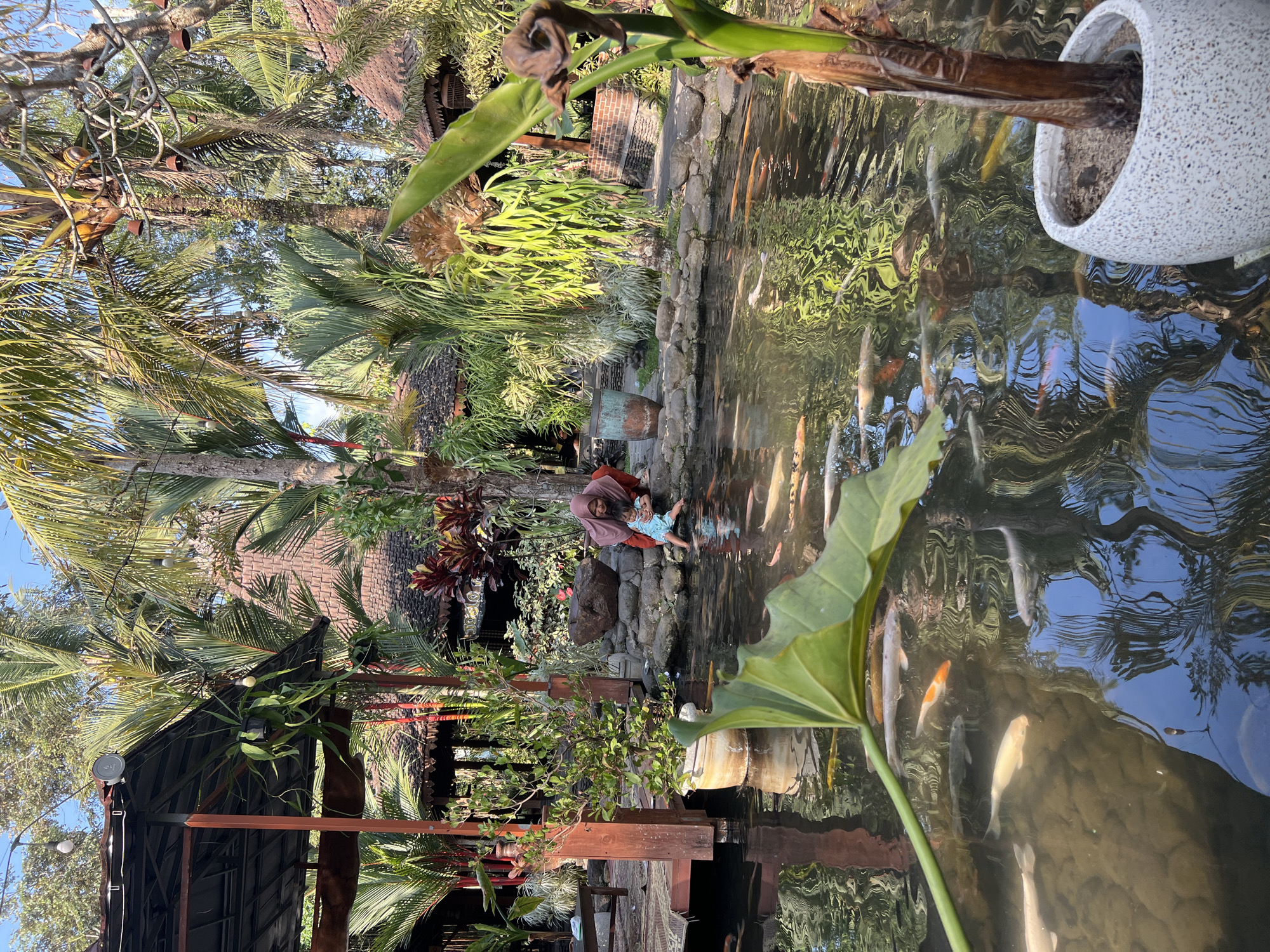 A woman wearing a maroon hijab and an orange shirt is kneeling by a pond filled with koi fish, holding a young child who is wearing a light blue outfit. The pond is surrounded by a rocky edge and lush greenery, including palm trees and various plants. In the background, traditional Indonesian architecture with thatched roofs is visible. The scene appears to be in a serene outdoor setting, likely a garden or resort, possibly located in Tlogoadi, Indonesia, given the architectural style and vegetation. The water of the pond reflects the surrounding trees and sky. In the foreground, a large green leaf from a plant is positioned close to the camera, partially obscuring the view of the pond. A potted plant with a brown trunk is also visible in the lower right corner of the image. The overall atmosphere is peaceful and familial.