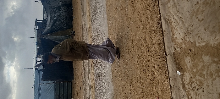 An older man with a white beard and dark cap is depicted walking from left to right across a muddy and sandy ground in Khan Yunis, Palestinian Territory. He wears a dark brown jacket with his left hand in a pocket, and light grey loose-fitting pants over dark sandals. In the background, under an overcast sky, are multiple makeshift shelters constructed from dark tarpaulins, some with white coverings, and patterned fabric sheets, supported by various poles. The immediate foreground shows wet, compacted earth transitioning to a drier, gravelly area.