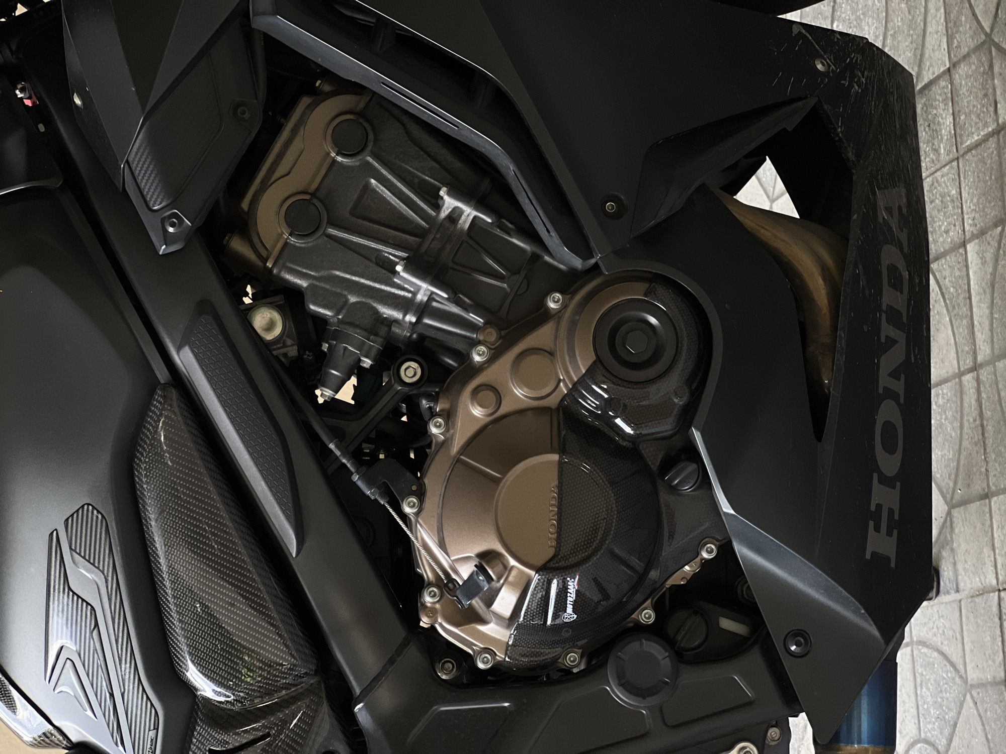 This is a close-up, eye-level shot of the engine and surrounding components of a black motorcycle. The lighting suggests it could be indoors or under overcast skies, as there are no strong shadows or direct sunlight.
The central subject is the motorcycle's engine, which features a mix of metallic finishes, including a bronze-colored clutch cover and silver-toned engine casings. A prominent carbon fiber accent is visible on the clutch cover, etched with the "HONDA" logo and another smaller logo that appears to read "SP TAKEGAWA." A black plastic fairing with a carbon fiber texture is also part of the engine cover. To the right of the engine, a portion of the black motorcycle bodywork is visible, also bearing the "HONDA" logo in a lighter gray color. The bodywork appears to have some dirt or scuff marks.
The rest of the image is filled with other parts of the motorcycle, including more black body panels, some of which have a textured surface. A blue exhaust pipe is partially visible at the bottom right. The background consists of a light-colored tiled surface, possibly a garage floor or patio.
No people are visible in the image, and no specific activity is taking place beyond the static presentation of the motorcycle's engine. The focus is on the intricate details and craftsmanship of the machinery.