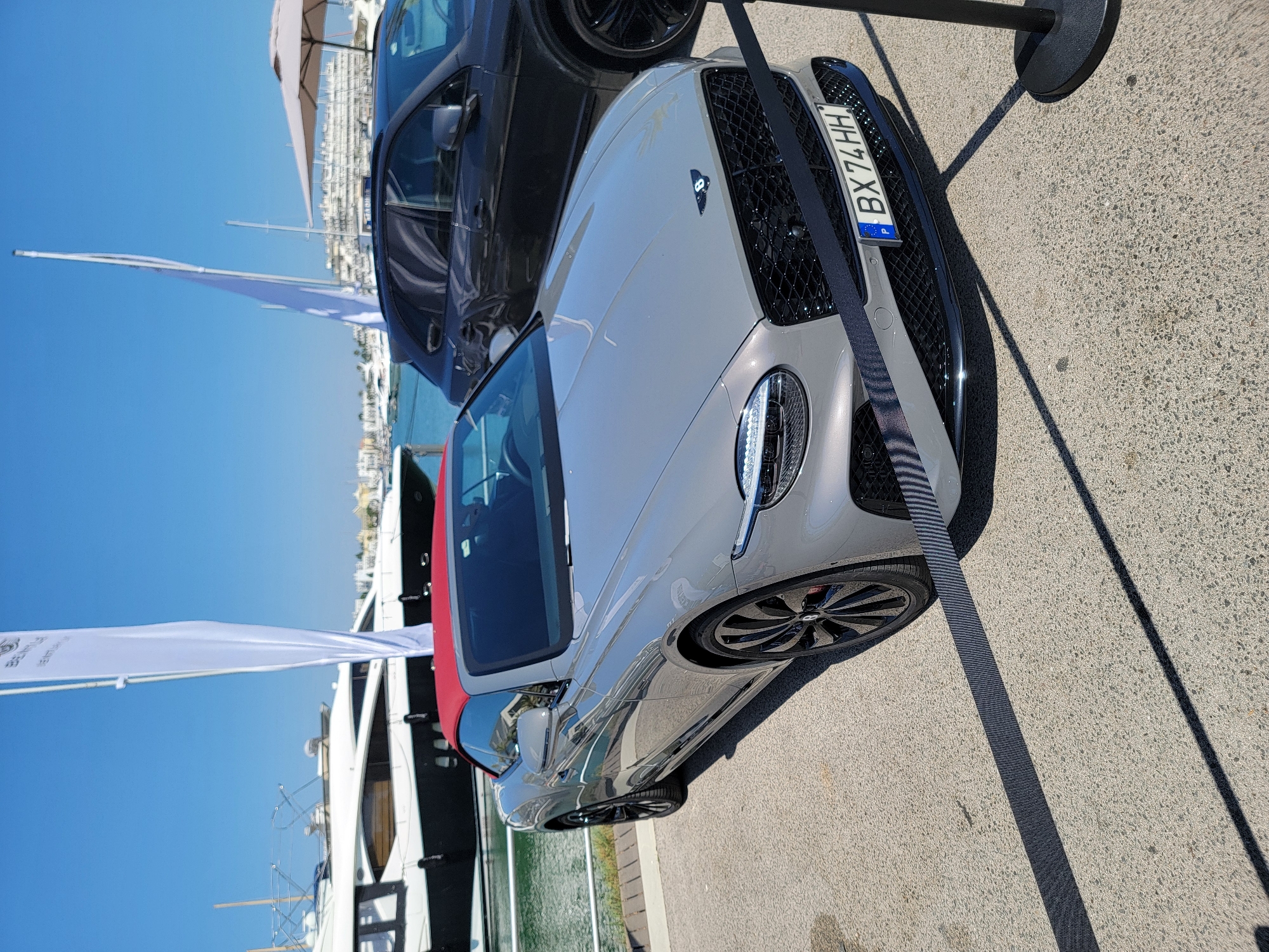 The image depicts a grey Bentley convertible with a red roof parked on a paved surface in Quarteira, Portugal. The car is positioned in the foreground, facing slightly to the left, with its license plate reading "BX 74 HH". Behind the Bentley, a dark SUV is visible, and further in the background, there are several yachts docked in a harbor under a clear blue sky. Two white flags with logos, possibly representing Bentley, are also present. The scene appears to be an outdoor event or exhibition in a marina area.