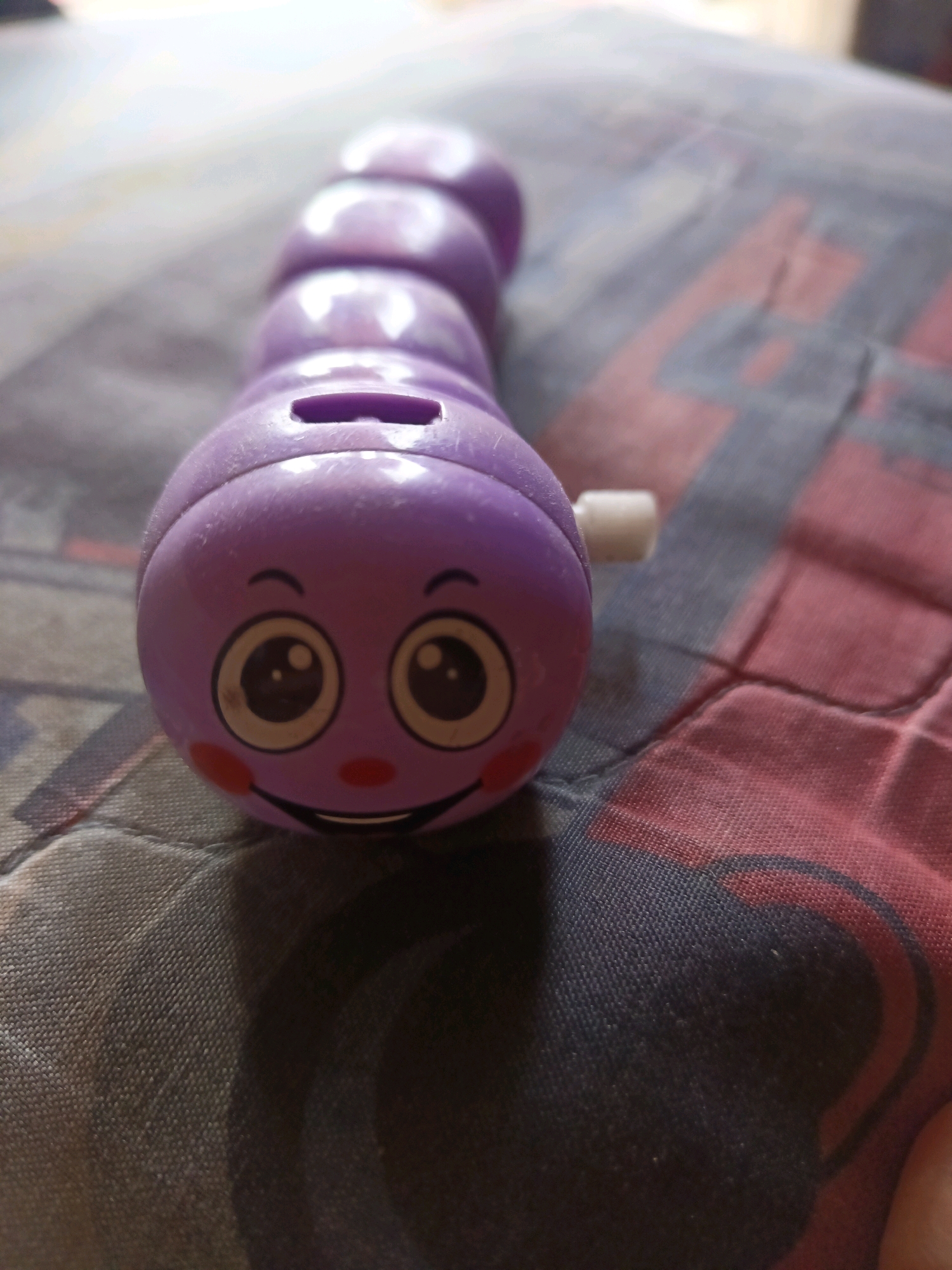 The image shows a close-up, eye-level shot of a purple, wind-up toy caterpillar lying on a patterned surface, possibly a blanket or tablecloth. The caterpillar is oriented horizontally, with its head facing left. The head is round and features a cartoonish face with large, black and white eyes, rosy cheeks, a red nose, and a smiling mouth. There is a slot on the top of its head, suggesting it might be a coin bank. A small white winding key protrudes from the right side of its head. The caterpillar's body is segmented, with several rounded sections extending to the right. The background surface has a pattern of interlocking, possibly geometric shapes in shades of dark blue, red, and gray, with some areas appearing lighter due to the lighting. The lighting in the image is somewhat dim, creating soft shadows and highlighting the glossy texture of the toy.
The location is stated to be in South Jakarta, Indonesia. This information is external to the visual content of the image itself.