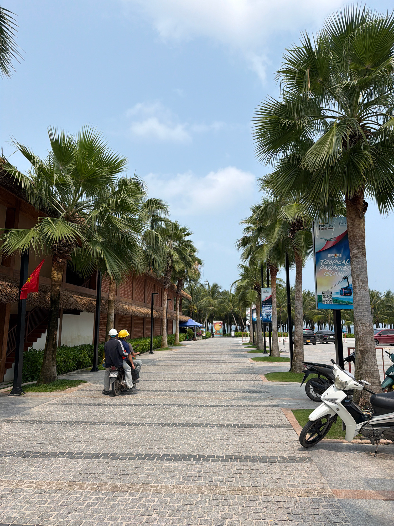 The image depicts a scene along a waterfront promenade in Phú Quốc, Vietnam, likely at a resort or tourist area. Tall palm trees line the path, casting shadows on the cobblestone walkway. Several motorcycles are parked along the side of the path. In the foreground, two individuals wearing hard hats ride a motorcycle, heading away from the viewer. To the left, a building with a thatched roof and a Vietnamese flag is visible. Further down the promenade, banners advertising "SUNWORLD TROPICAL PARADISE ISLAND" are displayed on palm trees, suggesting a commercial establishment. The sky is bright blue with scattered clouds, indicating fair weather. The overall atmosphere is that of a sunny day in a tropical destination.