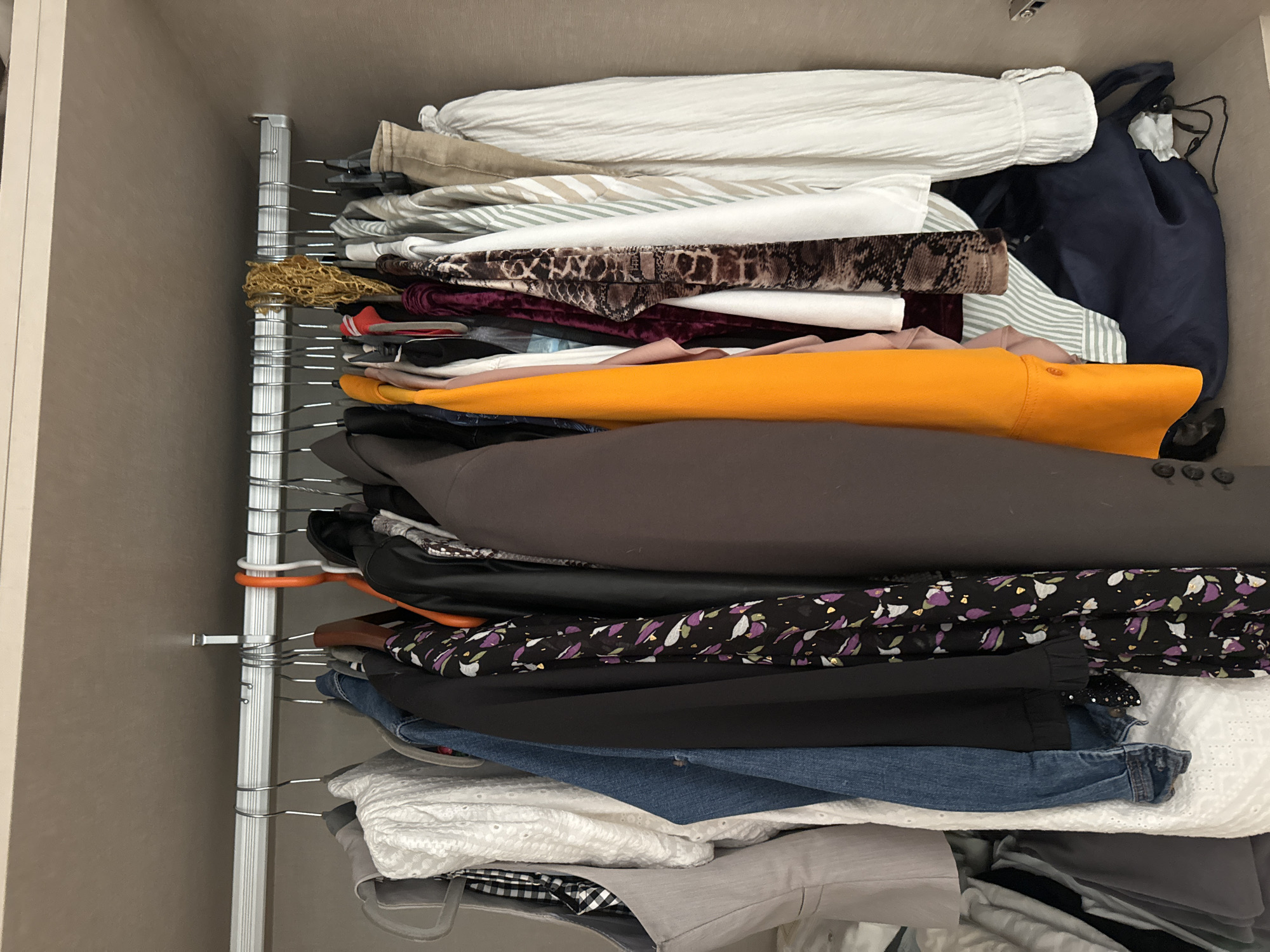 The image shows a close-up, eye-level view of an open closet. The closet is filled with various articles of clothing hanging on a metal rod. The clothing includes a grey vest, a white lace top, a denim jacket, a black jacket, a patterned dress, a bright orange top, a maroon velvet top, a snake-print top, and several striped and white shirts. A dark blue bag is visible at the bottom of the closet. The closet appears to be in a domestic setting, likely a bedroom. There are no people or animals in the image, and no specific landmarks or contextual clues that would indicate its location in Murcia, Spain.