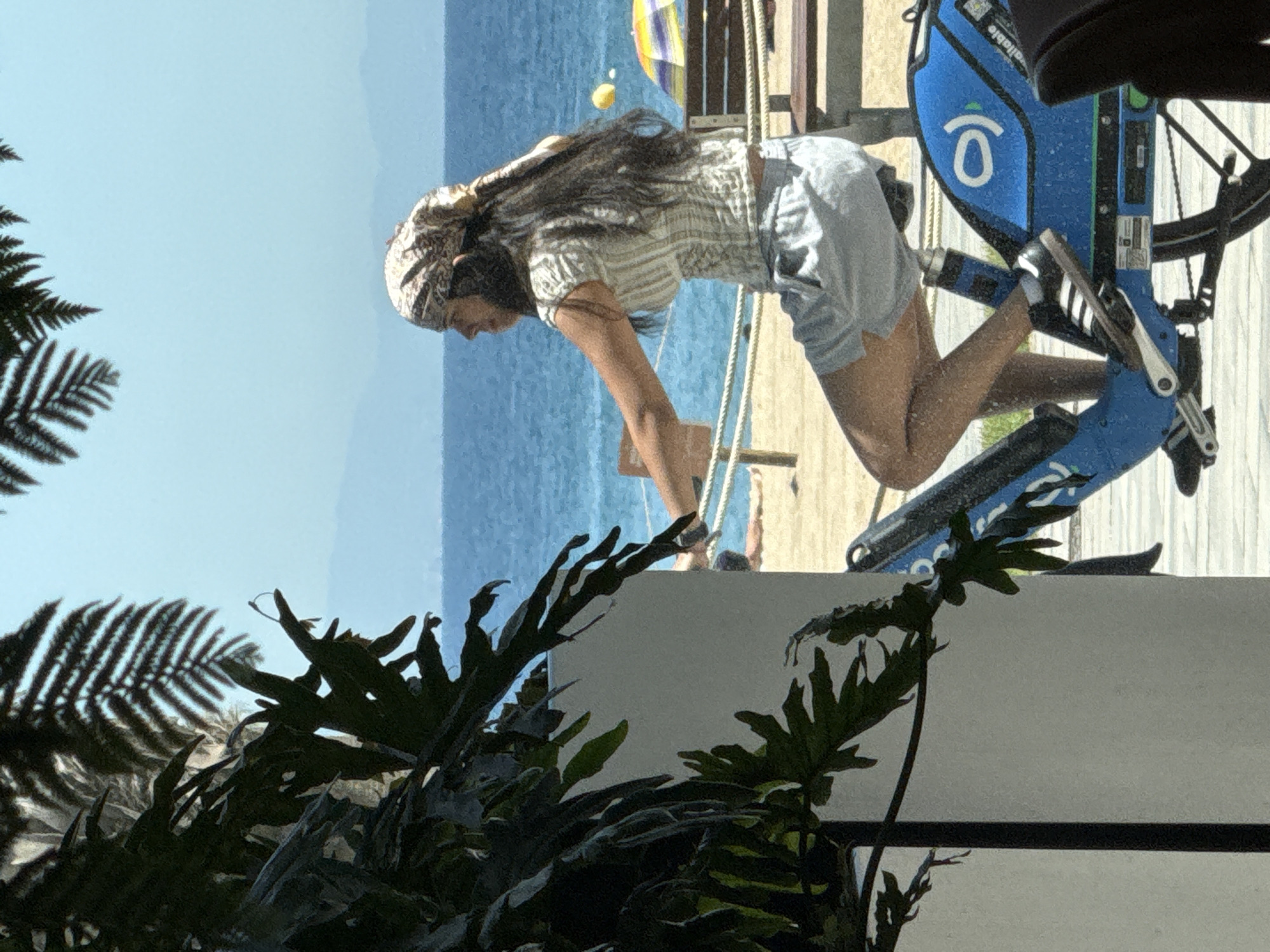 A woman with long, dark hair wears a patterned bandana and a striped shirt with light-colored shorts as she rides a blue bicycle with a white logo. She is on a paved surface, with a sandy beach and the ocean visible in the background. The ocean is a deep blue, and in the distance, faint mountains are visible under a clear sky. A yellow buoy floats on the water, and a colorful umbrella and a wooden bench are present on the beach. Lush green foliage frames the left side of the image, obscuring part of a white wall. The scene appears to be in Barcelona, Spain, likely near the waterfront.