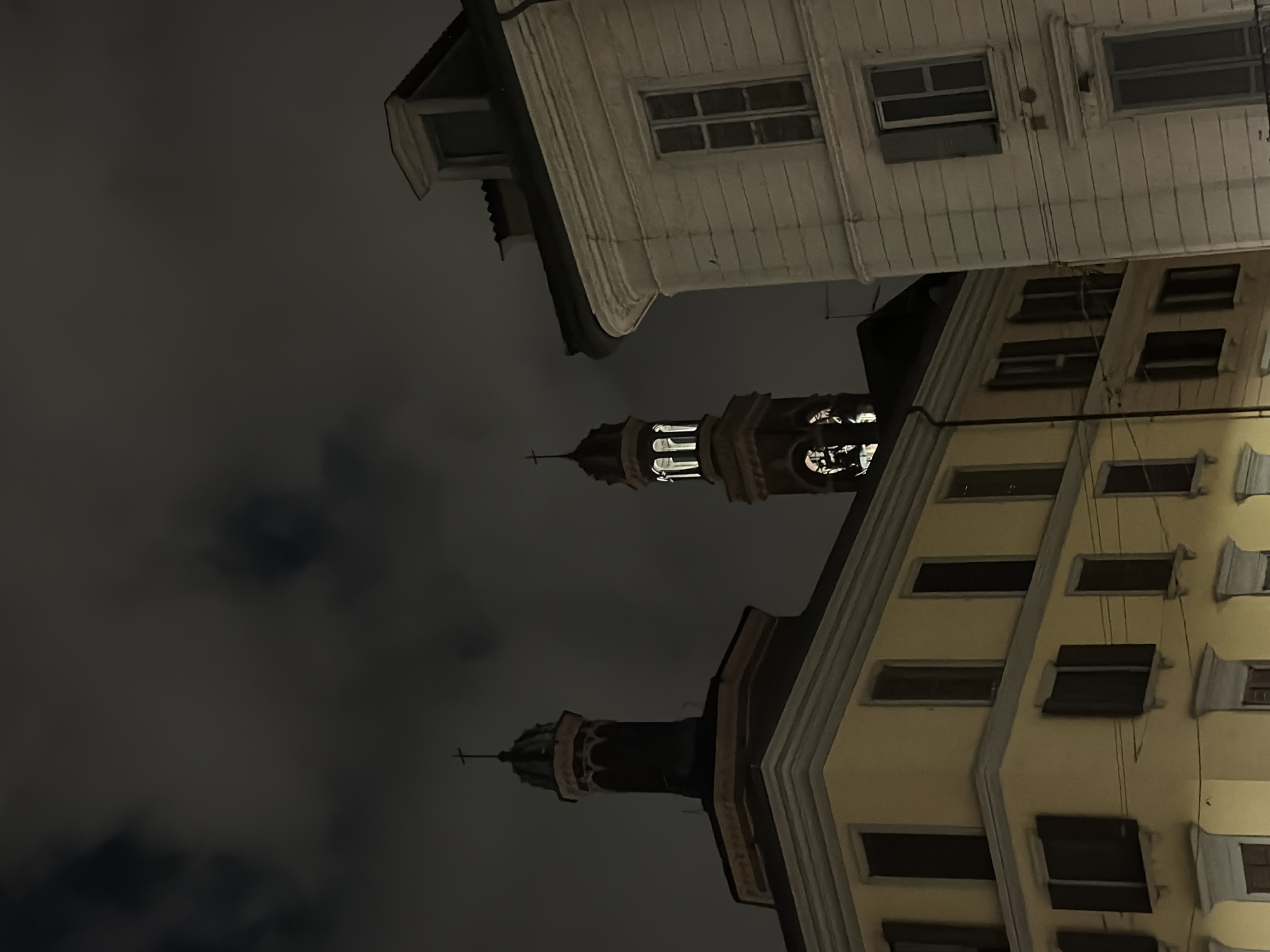 The image depicts an upward view of buildings and a church tower in Milan, Italy at night. The sky is dark and cloudy. Two buildings, one white and one yellow, are visible on the right side of the frame. The buildings appear to be multi-storied, with windows and shutters. A church tower with a cross on top is positioned between the buildings. The tower is lit up, revealing architectural details. Wires are visible running along the facade of the yellow building.