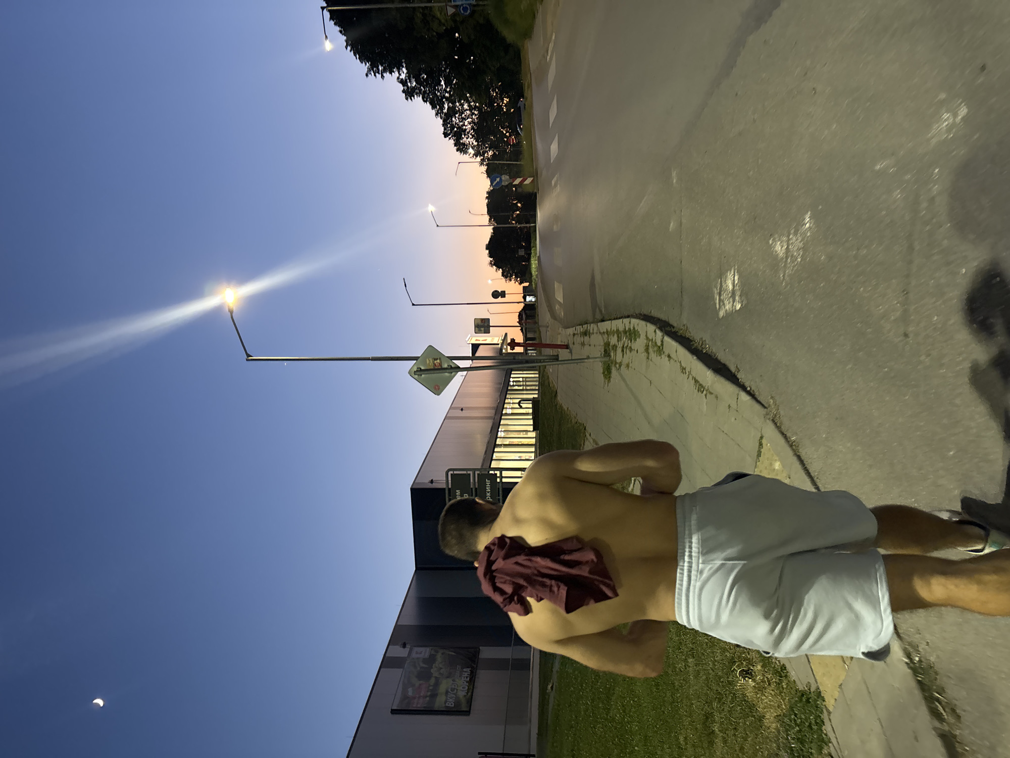 A shirtless person with a maroon towel draped over their shoulders walks along a paved sidewalk next to a road. The person is wearing light gray shorts and white sneakers. The sun is setting, casting a warm orange glow on the horizon. Streetlights are on, and the sky is a deep blue with a crescent moon visible in the upper left corner. To the left of the sidewalk is a grassy area and a large, modern building with a sign that appears to be in Cyrillic script. The road curves to the right and ascends a slight incline. The scene suggests a suburban or edge-of-city location at dusk.