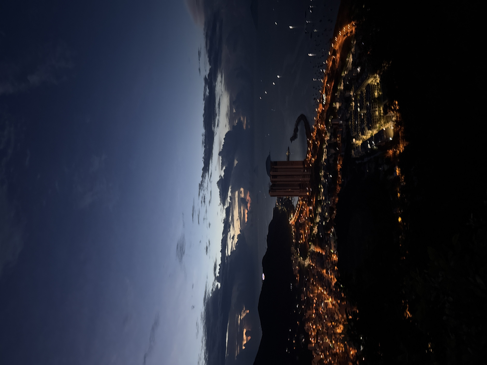 The image shows an aerial view of Bac Nha Trang Ward, Vietnam at dusk or night. In the middle of the image is a tall, rectangular building, likely a hotel or apartment complex, illuminated from within. The city stretches to the right of the building, a patchwork of residential and commercial areas illuminated by streetlights and building lights. In the foreground is a dark, hilly area, potentially a park or natural reserve, partially obscuring the cityscape. To the left of the image, the sky transitions from a deep blue to lighter hues, with visible clouds and hints of a setting sun. The sky reflects the transition from day to night, with darker tones dominating the right side, suggesting that night has already fallen in that area.