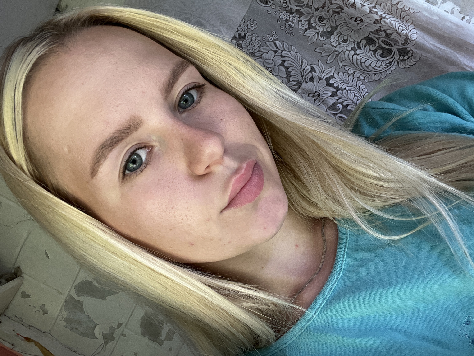 This is a close-up, horizontal, selfie-style photograph of a young woman with blonde hair. She is looking towards the camera with a neutral expression. Her light blue eyes are clearly visible, and she has fair skin with some freckles and minor blemishes. Her lips are slightly parted and appear to be colored with a light pink lipstick. Her blonde hair is long and parted to her right, with strands falling across her face and shoulders.
She is wearing a turquoise crew-neck t-shirt. A thin, silver-colored chain necklace is visible around her neck. The background is blurred, but appears to be a patterned fabric, possibly a tablecloth or bedding, with a floral design in shades of gray and white. To the left of the frame, there is a section of a wall with peeling paint, suggesting an indoor setting.
The lighting appears to be natural and soft, suggesting it might be daytime, but there are no strong shadows or highlights to indicate a specific time of day or weather conditions. The overall mood of the image is relaxed and informal. There is no visible text in the image.