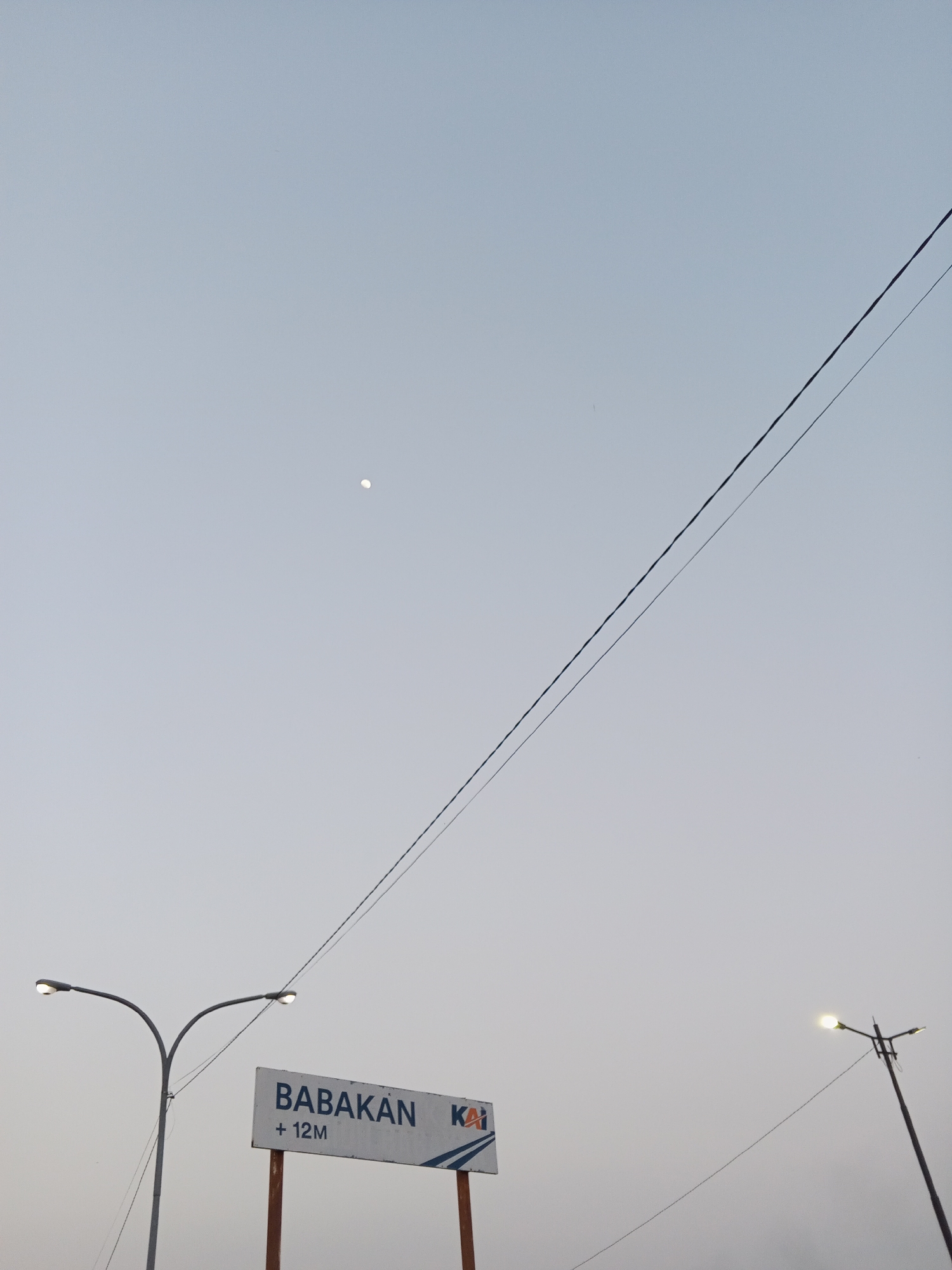 A street sign for "BABAKAN +12M KAI" stands in the foreground, supported by two wooden posts. The sign features blue and white lettering. To the left of the sign, a double-headed street lamp with illuminated lights is visible. To the right and further back, another street lamp with multiple lights is also illuminated. Overhead, two parallel power lines stretch across the sky, angled from the lower left to the upper right. The sky is a pale blue-grey, with a small, faint circle, possibly the moon, visible in the upper left quadrant. The overall scene suggests an outdoor setting, likely near a railway, given the "KAI" marking on the sign. The lighting indicates either dawn or dusk. The location is identified as Pakusamben, Indonesia.