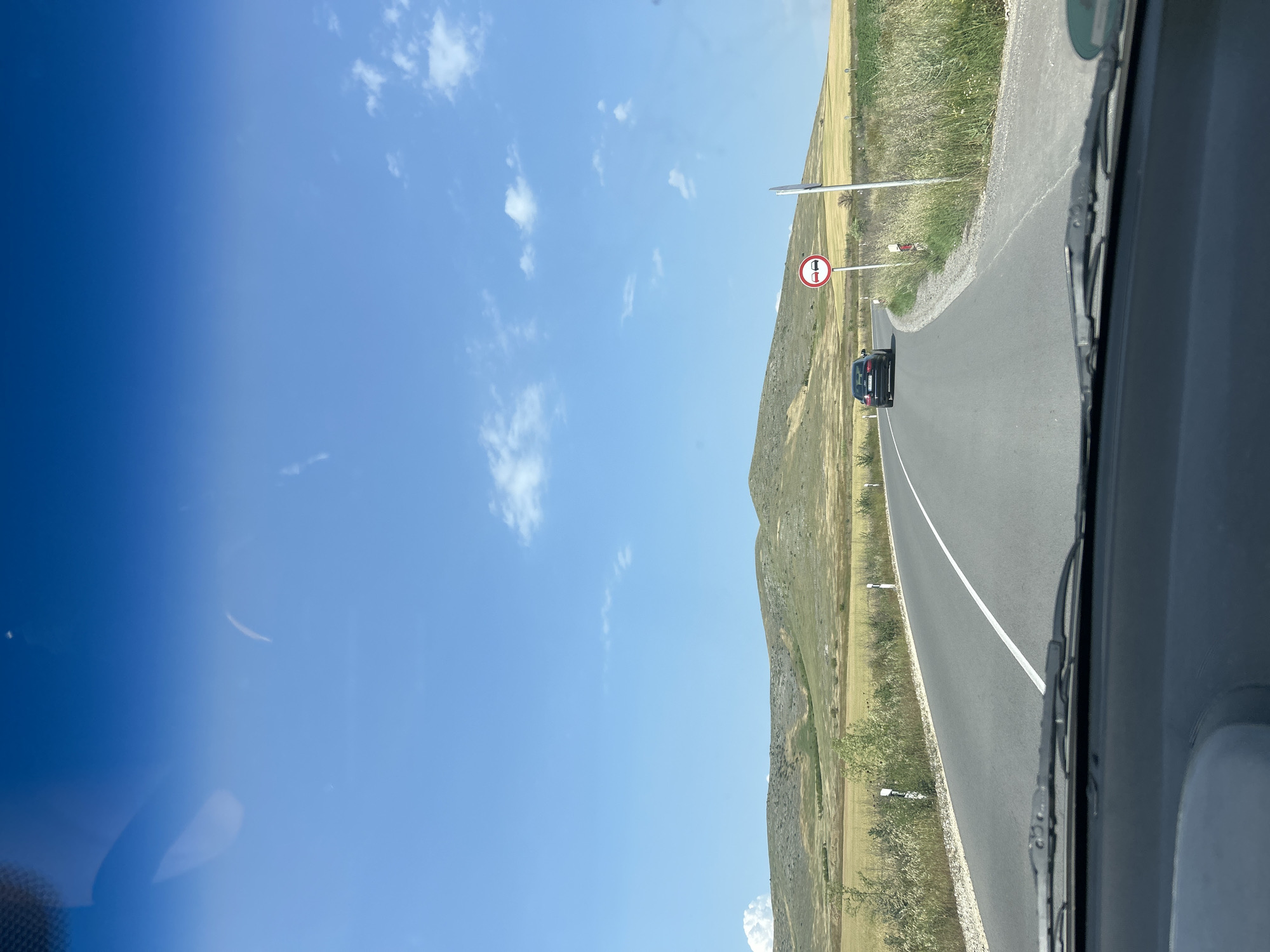 The image depicts a daytime scene from the interior of a vehicle, looking forward through the windshield, with the dashboard and wipers visible in the foreground. The location is Isperihovo, Bulgaria. A two-lane asphalt road, marked with a solid white center line, curves gently to the right. Ahead, a dark-colored sedan, possibly an Audi, travels in the same direction. On the right side of the road, a circular "No Overtaking" regulatory sign, featuring two cars crossed out in red, is mounted on a pole, accompanied by a smaller white delineator post. The landscape beyond the road consists of rolling hills covered in a mix of dry, yellowish-brown grass and green vegetation, extending towards the horizon. Sparse white clouds are scattered across a clear, bright blue sky.