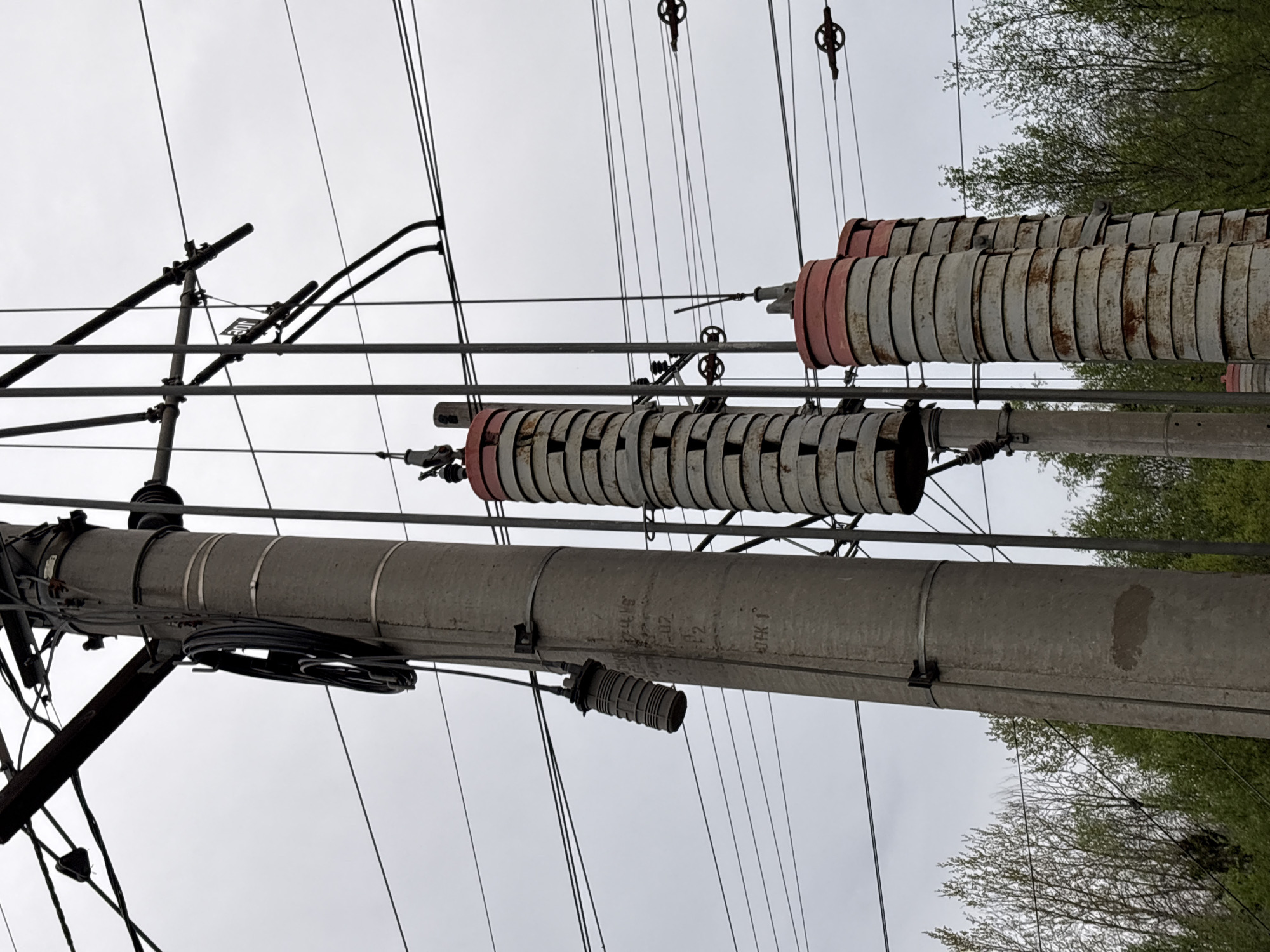 The image shows a utility pole with various electrical components against a cloudy sky and some trees on the right side of the frame. The utility pole is concrete and has several wires and cylindrical devices attached to it. There are metal rings or disks stacked on some of the cylinders, with some having red end caps. Various wires and support structures are visible above and around the pole. The environment is outdoors, likely in an urban or suburban area. The image's metadata states it was taken in Solnechnoye, Russia.