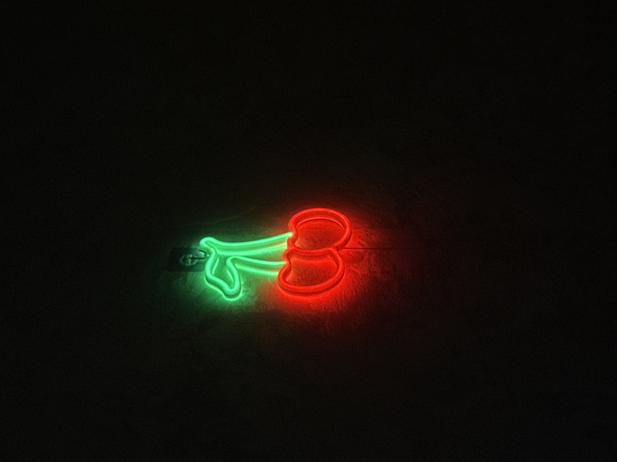 The image shows a neon sign shaped like two cherries on a dark, textured wall. The cherries are depicted in red neon, and the stem connecting them and a small leaf are in green neon. The lighting is dim, with the neon lights being the primary light source, casting a glow on the wall. The location of Misaylovo, Russia is not visible or implied in the image.