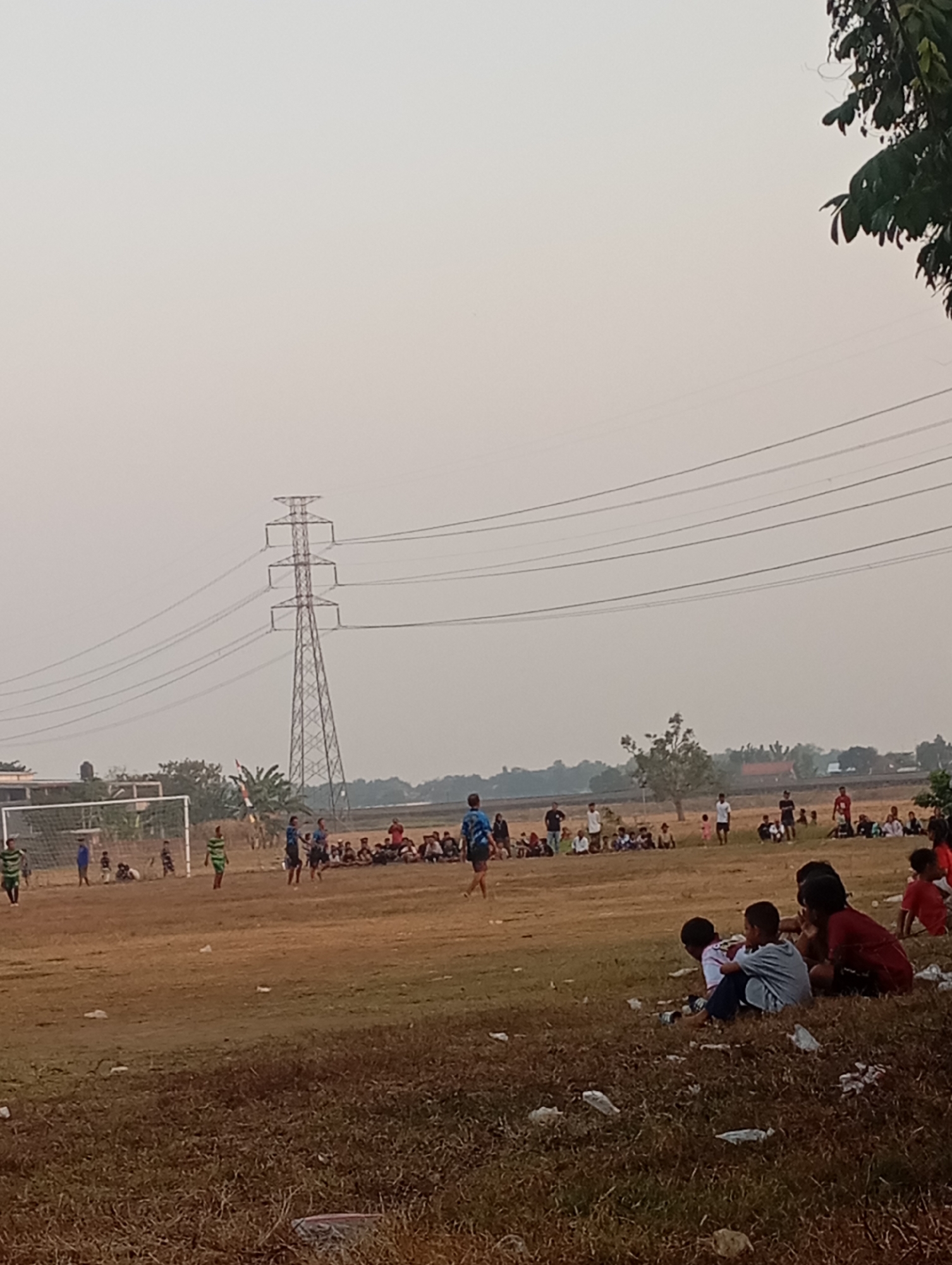 In Kecamatan Haurgeulis, Indonesia, a football match is underway on a dry, brown field under an overcast sky. A large, lattice-style electricity pylon stands prominently in the background, with power lines stretching across the horizon. Spectators, mostly children and adults, are seated and standing along the sidelines, some watching the game and others gathered in groups. A goalpost is visible on the left side of the field. Several children are playing on the field, while others are seated in the foreground, some appearing to be resting or observing. The environment is rural, characterized by sparse vegetation and a hazy atmosphere.