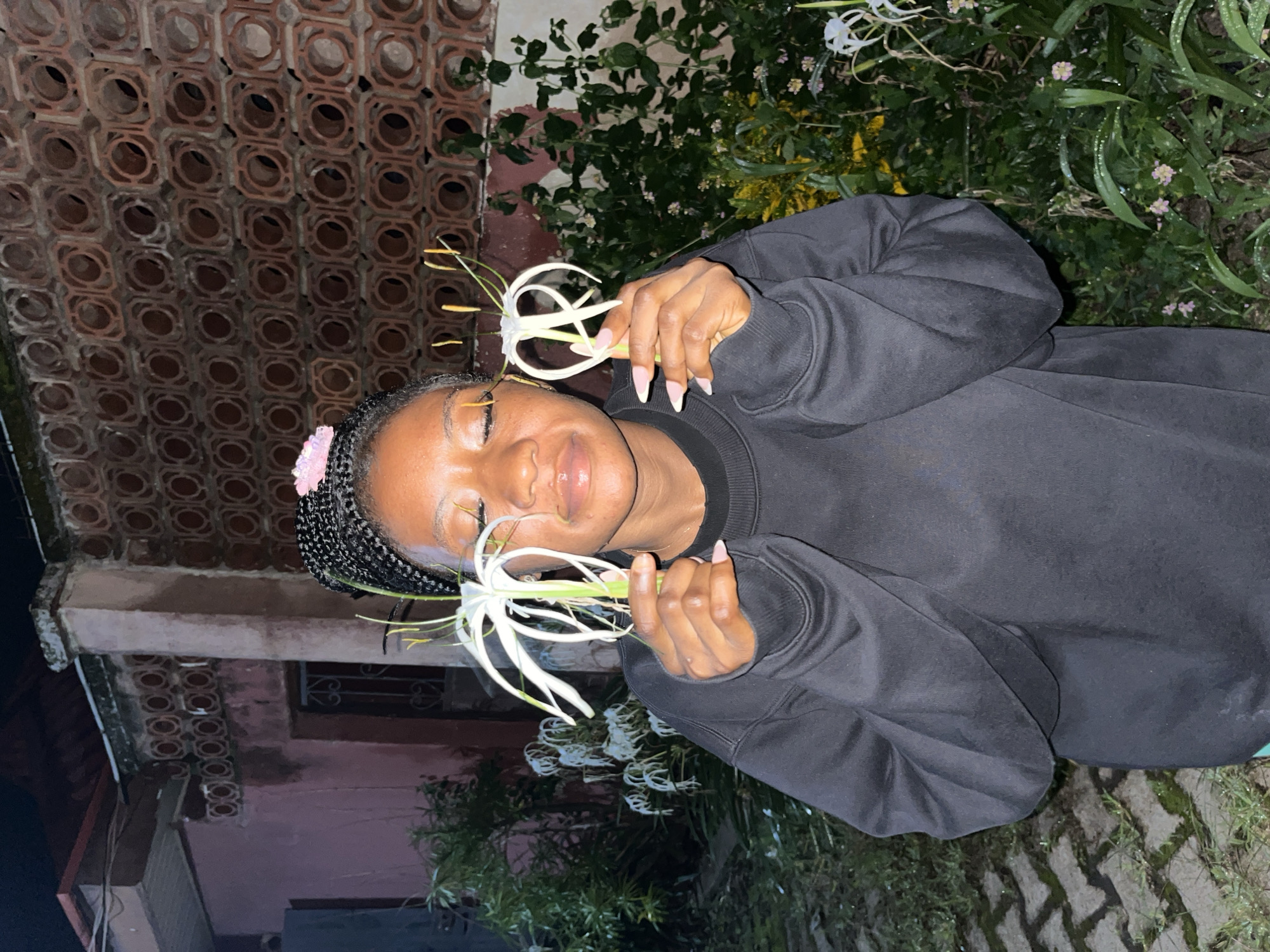 A young Black woman with braided hair, adorned with a pink clip, stands outdoors at night, holding two white spider lilies. She is wearing a black crewneck sweatshirt and has a light pink manicure. Her eyes are closed, and her head is tilted slightly. The background features a textured brick wall, foliage, and what appears to be a building. The ground is paved with interlocking stones and is wet, suggesting recent rain. The scene is located in Jos, Nigeria.