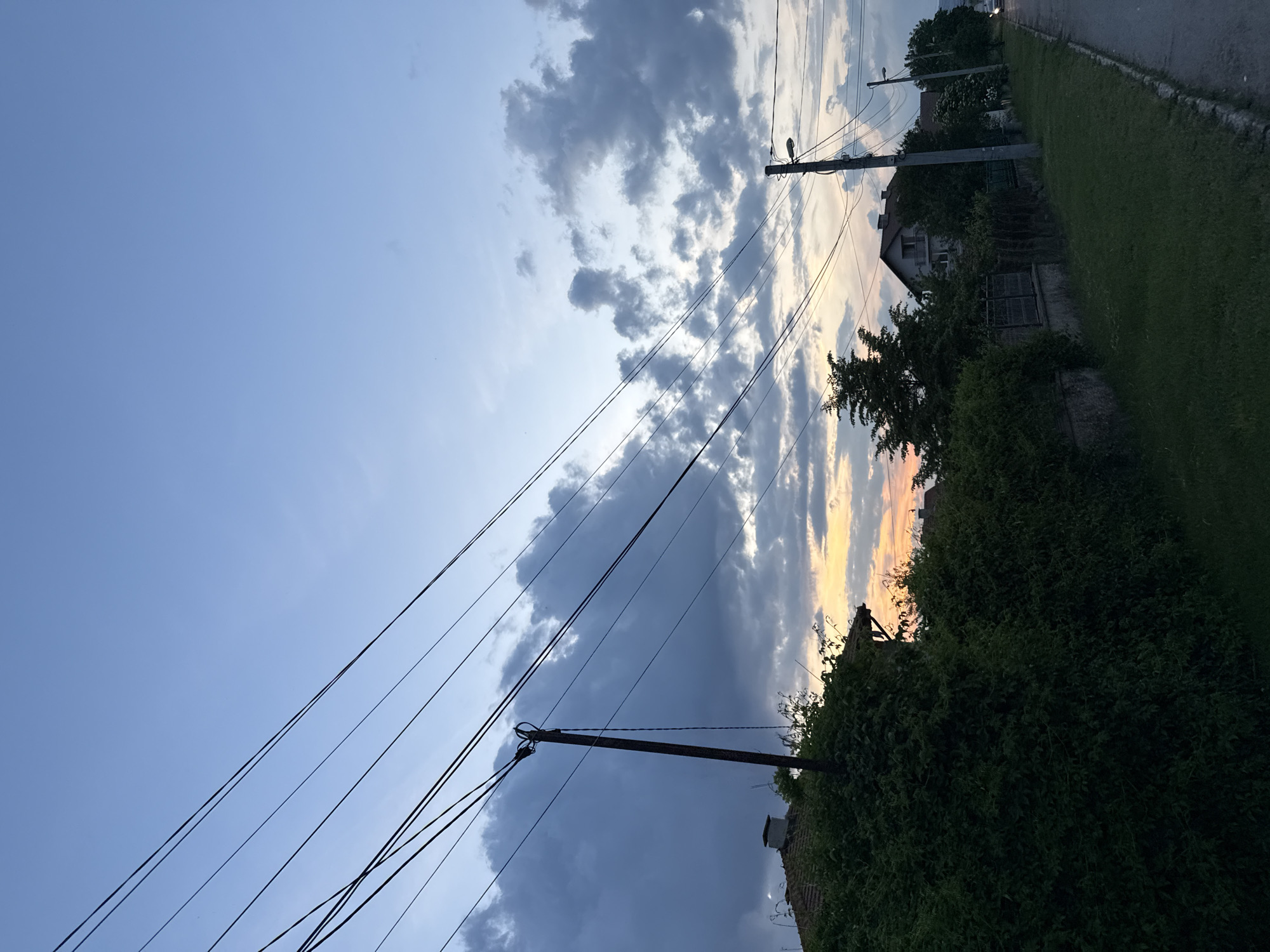 This image captures a residential street scene in Mirovyane, Bulgaria, at dusk. The sky transitions from a clear, pale blue at the upper left to an array of grey and white stratocumulus clouds, with distinct warm orange and yellow light from the setting sun visible along the lower horizon. Multiple dark utility lines diagonally cross the upper portion of the frame. In the mid-left, a prominent dark utility pole stands, supporting several power lines, with one cable hanging freely. Below the pole, the left foreground is dominated by a dense, dark green hedge or overgrown vegetation, partially obscuring a building with a tiled roof and a chimney. To the right, a paved road stretches into the distance, bordered by a narrow grass verge and a low stone wall topped with a dark metal fence. Beyond this, further trees and a multi-story house with a grey roof and light-colored walls are visible. Another utility pole equipped with a street light stands further along the road on the right. In the far distance on the road, faint vehicle headlights are discernible. No individuals are present in the visible frame.