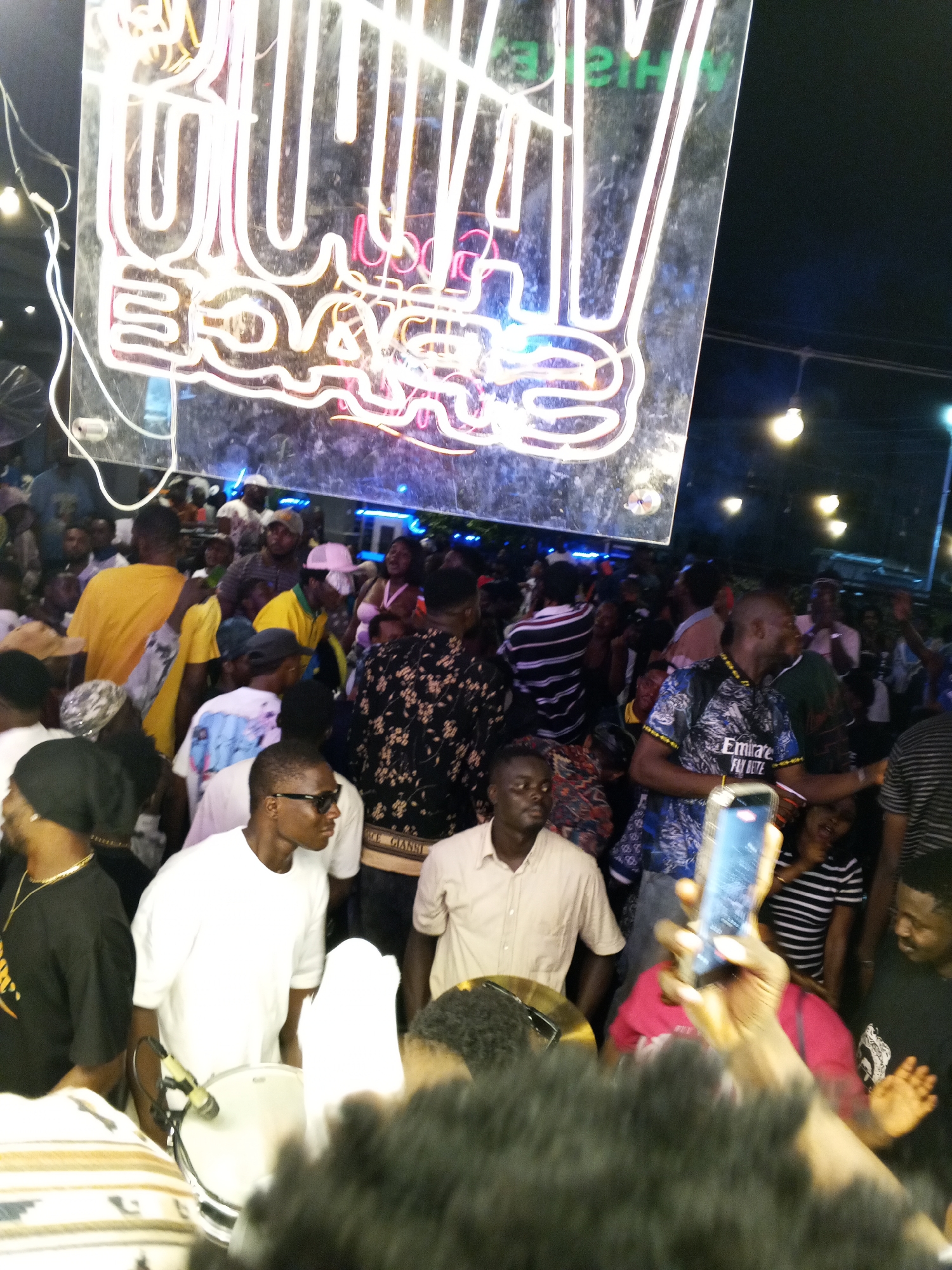 The image depicts a crowded scene at night, likely in Lokoja, Nigeria. A large neon sign with the words "ROYAL BOYS" illuminated with white and pink lights hangs overhead. The crowd is composed of many people of African descent wearing various clothing styles. A man in a white shirt and sunglasses stands near a drum. Another man in a light brown shirt stands beside him. Someone in the foreground holds up a smartphone, potentially recording the event. Streetlights are visible in the background.
