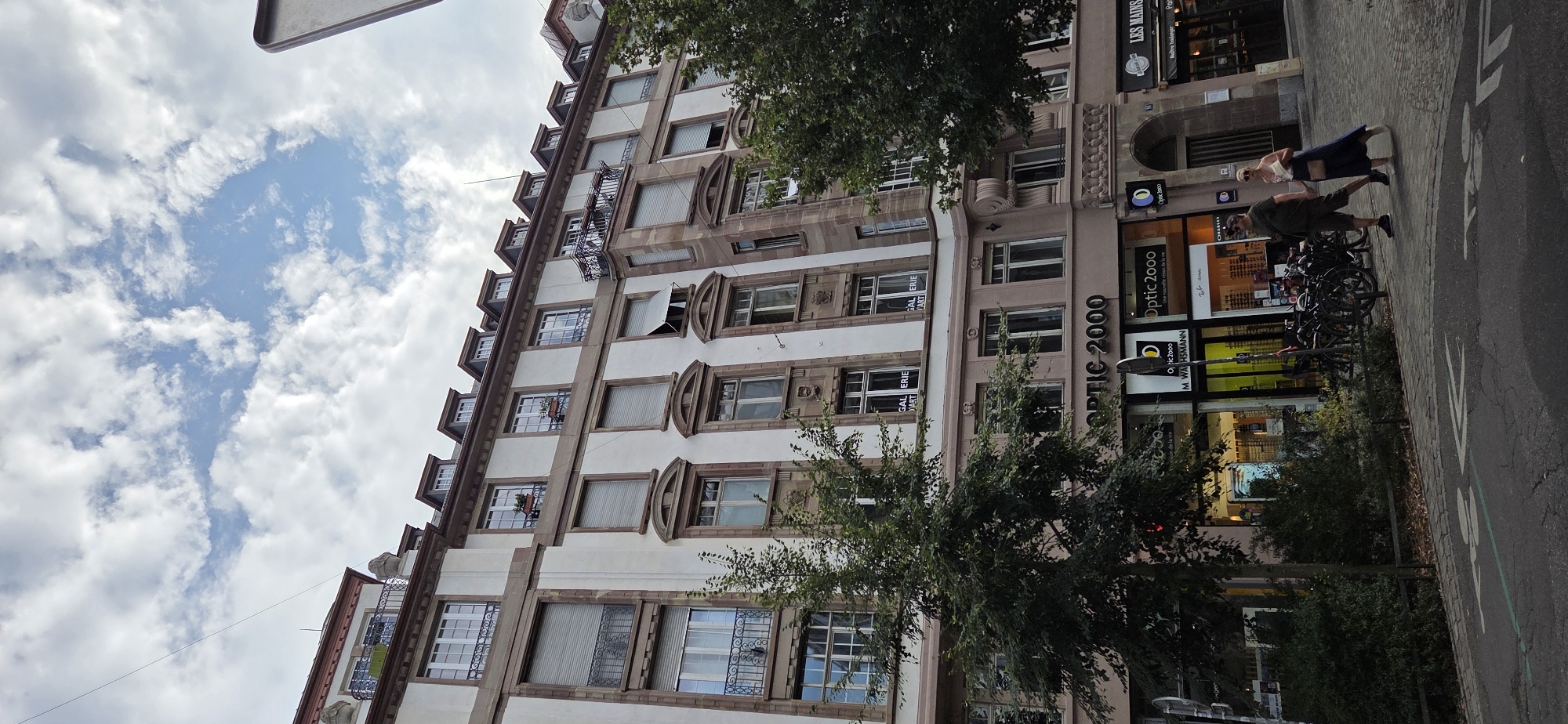 The image shows a street view in Strasbourg, France. A multi-story building with a white facade and brown architectural details dominates the scene. The building has numerous windows, some with closed shutters and some open. Signs for "OPTIC 2000" and "LES MAINS" are visible on the ground floor. A tree stands in front of the building, partially obscuring the lower floors. Two people, a man and a woman, are walking hand-in-hand on the sidewalk in front of the shops. Bicycles are parked along the sidewalk. The sky is partly cloudy with a mix of blue sky and white clouds. The street has a designated bicycle lane marked with white arrows.